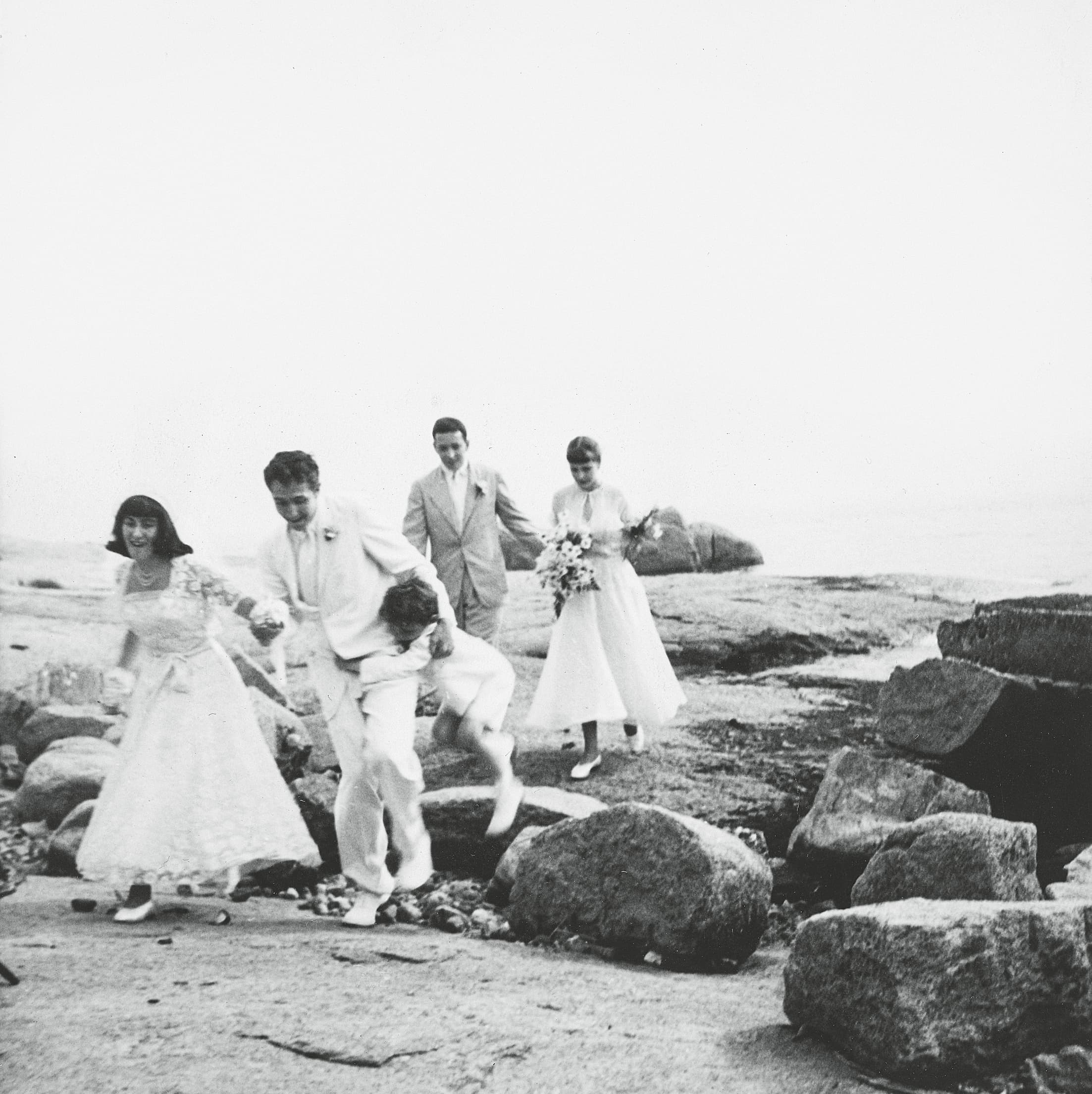 A newly married couple and two young girls playfully run along a rocky shoreline during a bright, sunny day.