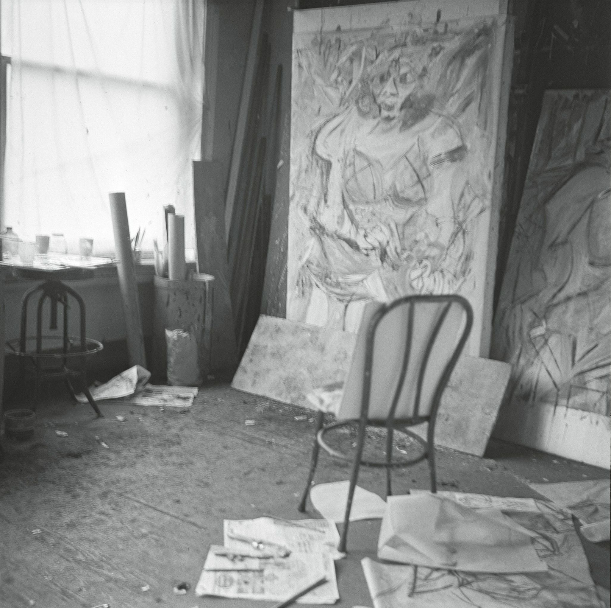 A cluttered artist's studio featuring a wooden chair in the foreground, scattered papers and art supplies on the floor, and large, unfinished paintings on the walls.
