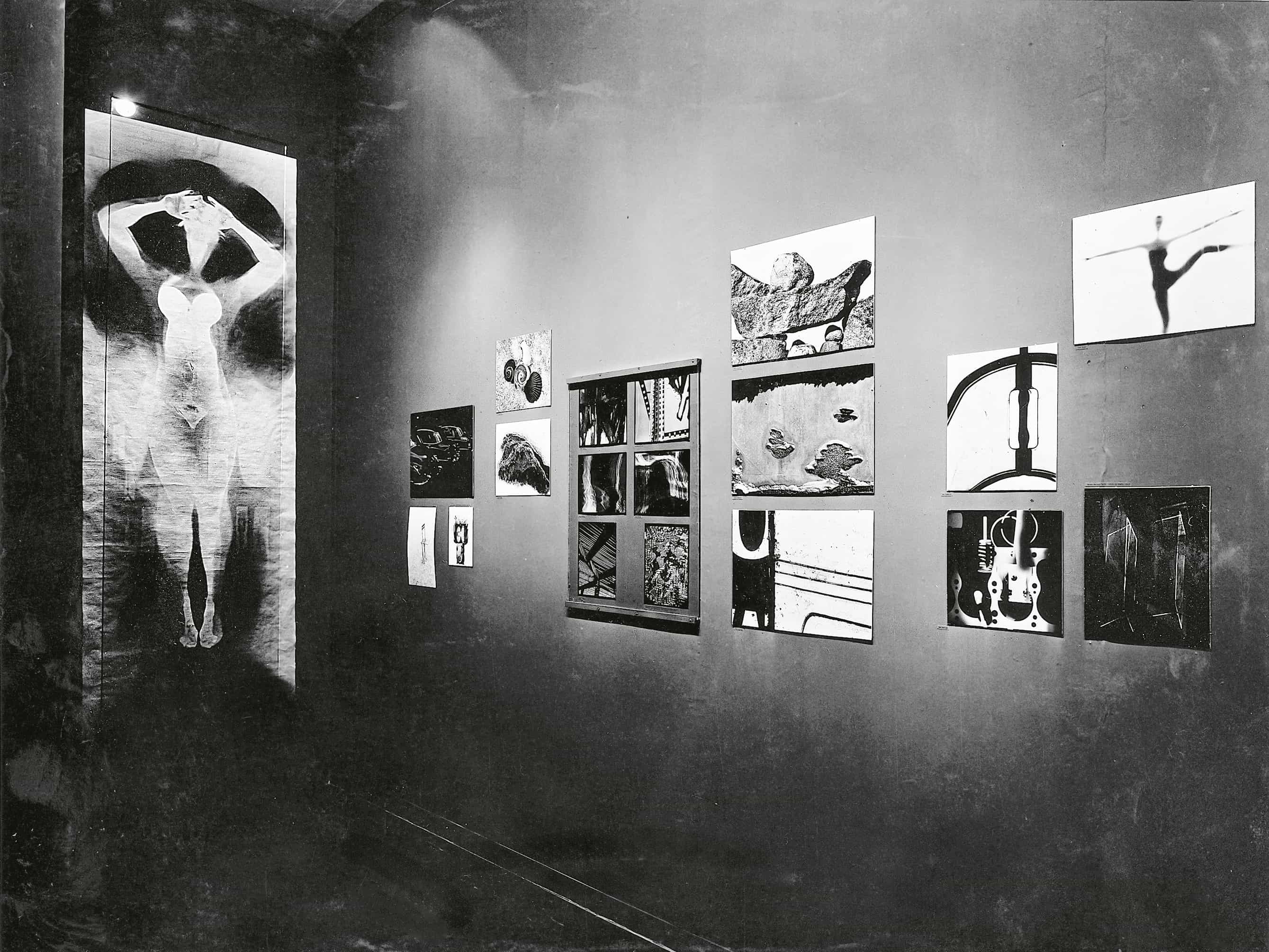 A black and white photograph depicts a gallery wall displaying a collection of abstract and surrealist artworks.