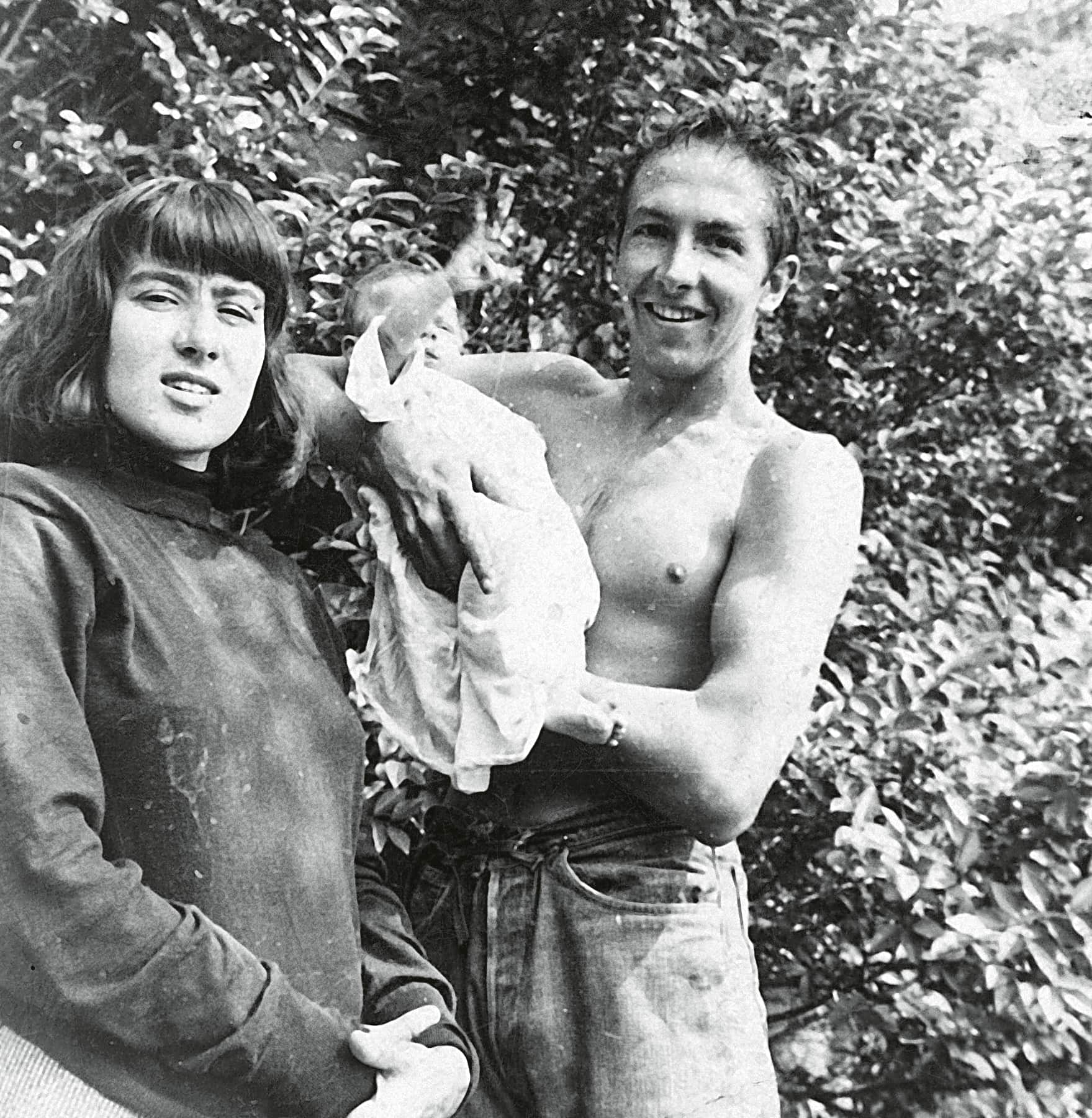 A smiling man holds his newborn baby outdoors amidst lush greenery.