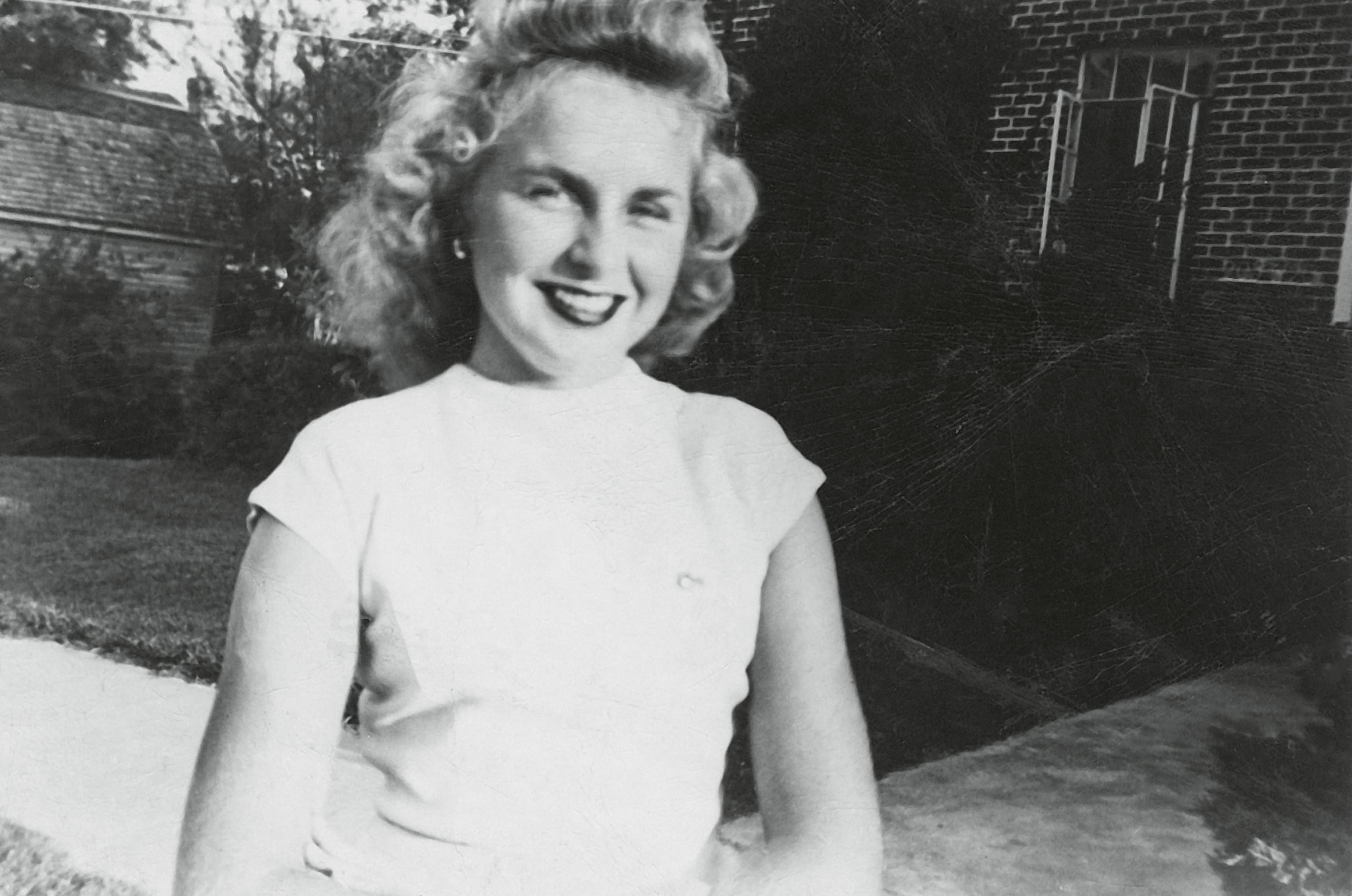 A smiling woman with curly hair is sitting outdoors, wearing a light-colored top, with a brick building and greenery in the background.
