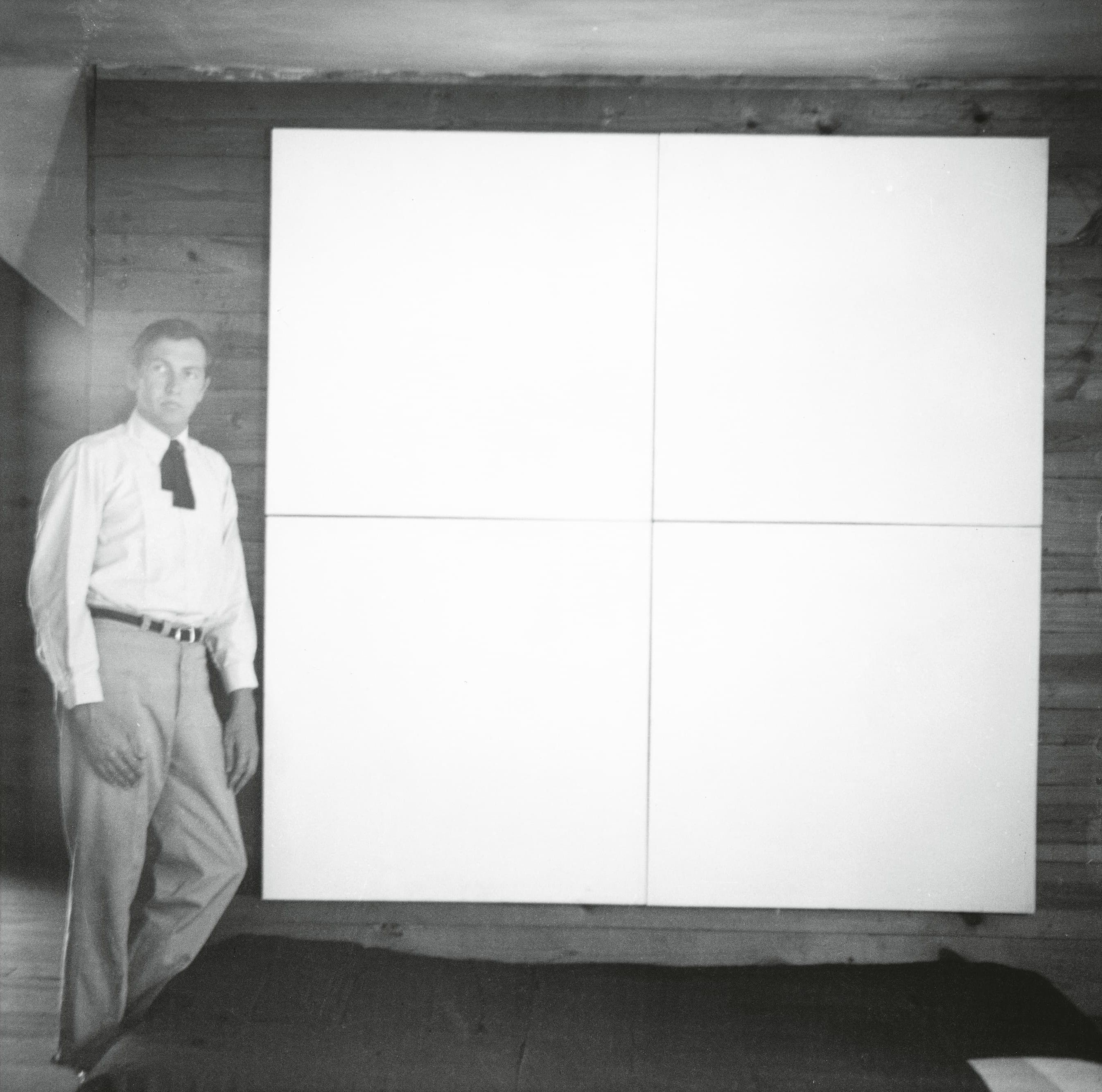 A man in a white shirt and tie stands beside a large, blank, four-panel white canvas mounted on a wooden wall.