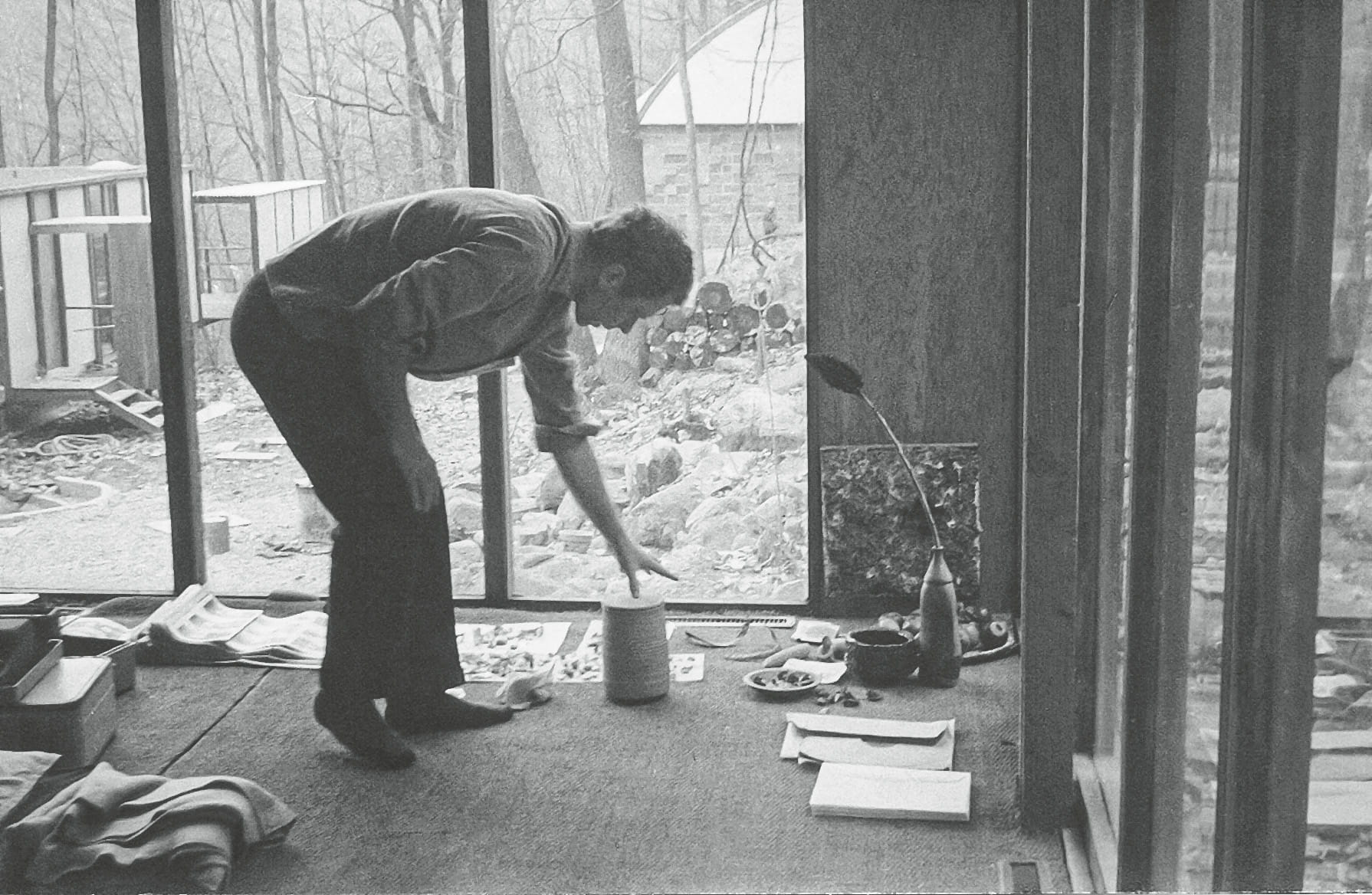 A man is bending over to place an object on the floor inside a room with large windows, revealing a view of a wooded area outside, while various items are scattered around him.