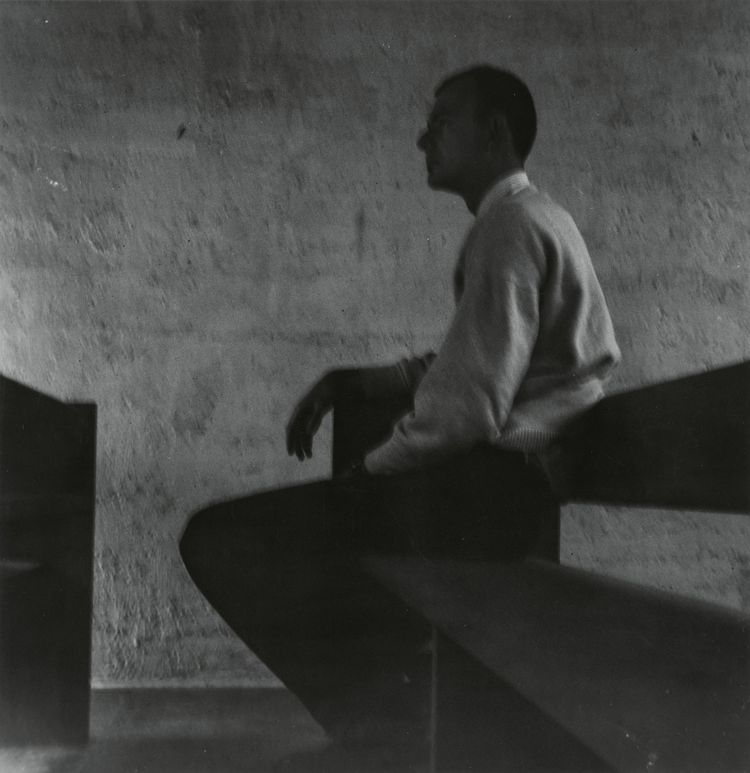 A grayscale photograph depicts a man sitting on a curved bench in a room with a textured wall, creating a sense of solitude and contemplation.