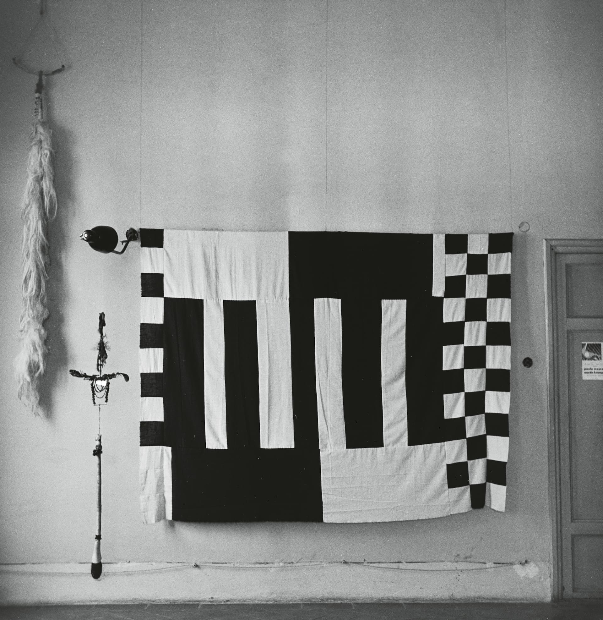 A black and white quilt featuring geometric patterns hangs on a wall, accompanied by a decorative staff and a hanging piece of fabric.