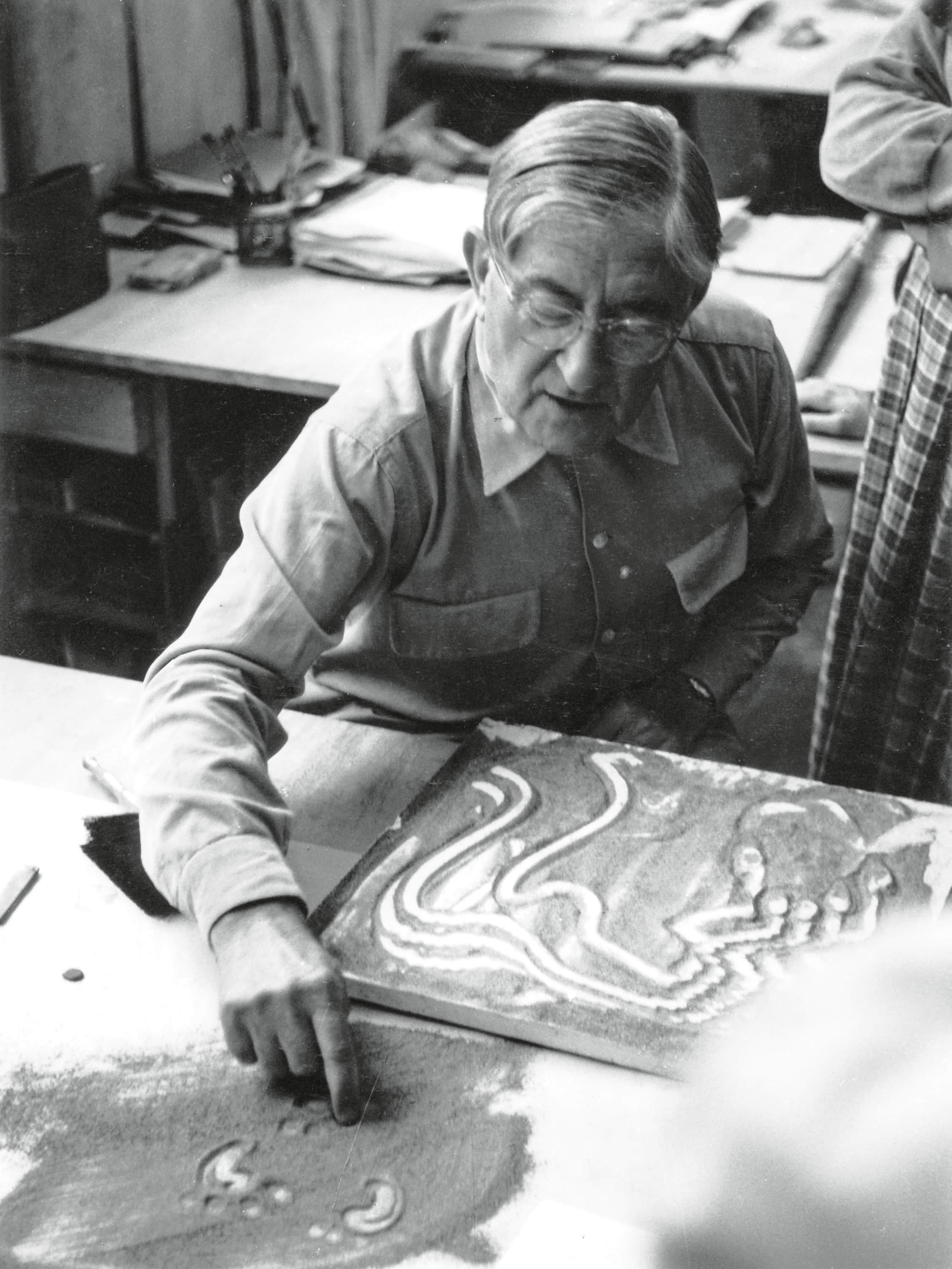 A man with glasses intently works on a drawing of a snake on a large, light-colored surface.