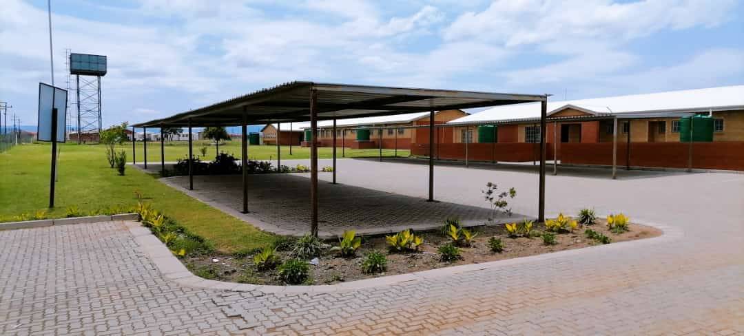 A long carport with a corrugated roof covers a paved area, with single-story buildings and a water tower in the background.