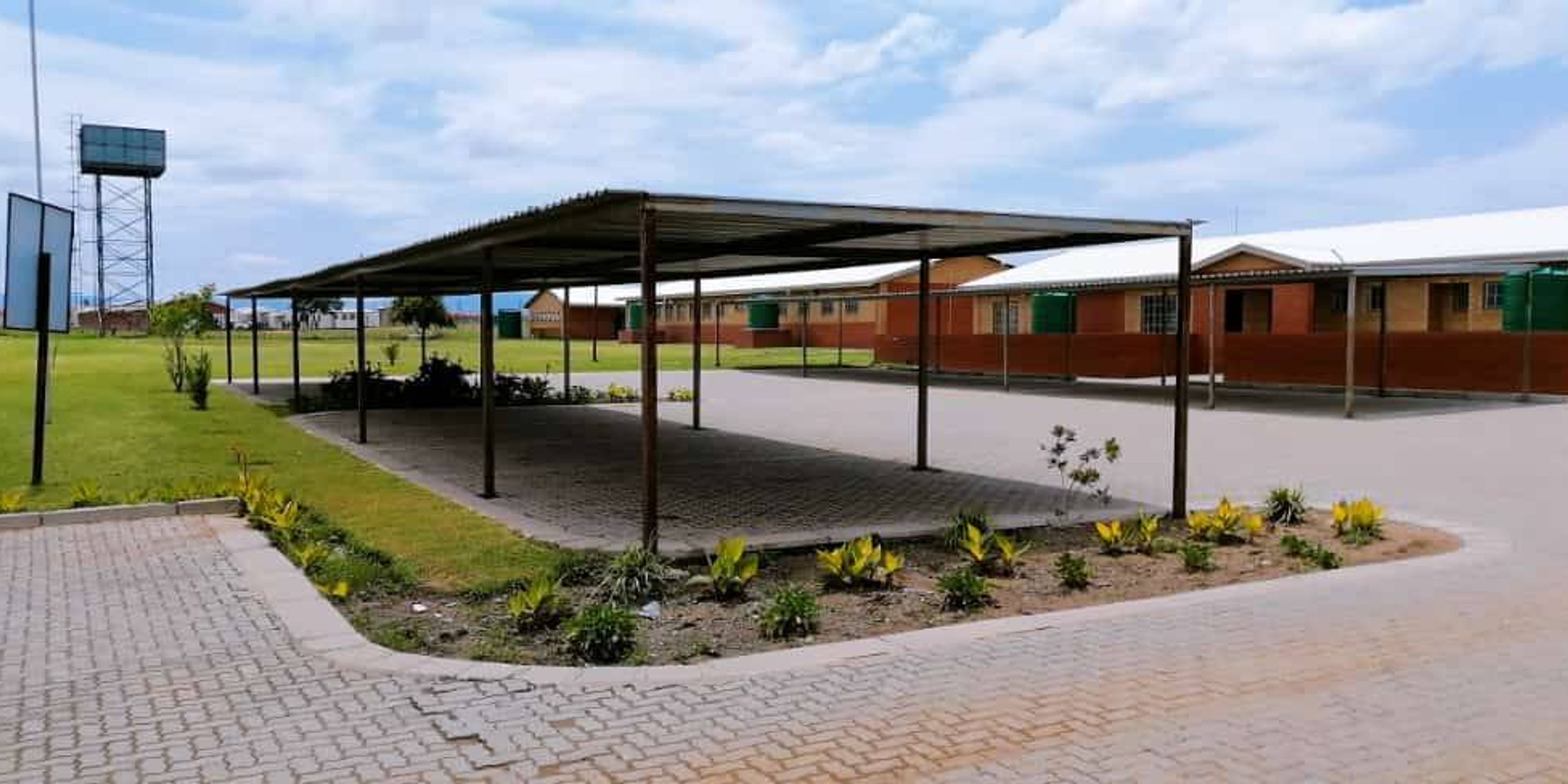 A long carport with a corrugated roof covers a paved area, with single-story buildings and a water tower in the background.