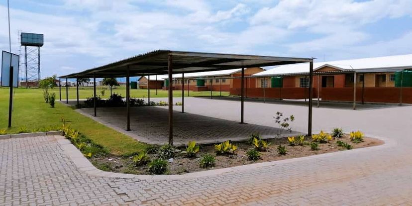 A long carport with a corrugated roof covers a paved area, with single-story buildings and a water tower in the background.