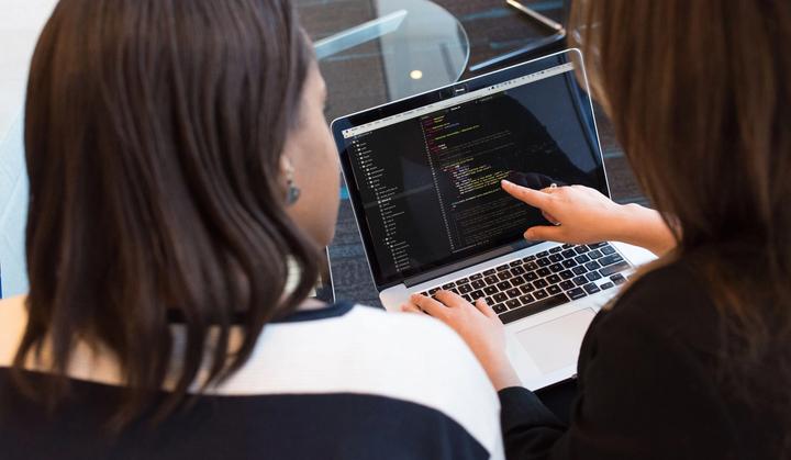 Two women look at code on a laptop together.