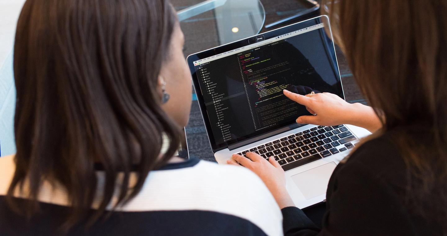 Two women look at code on a laptop together.