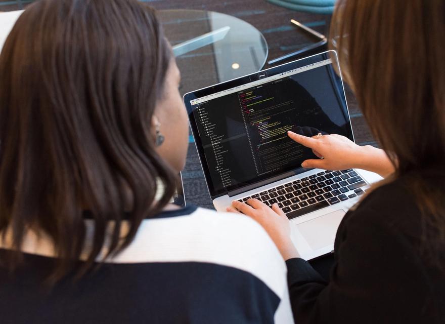 Two women look at code on a laptop together.