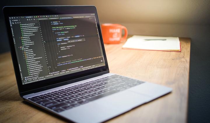 A laptop displaying code sits beside a notebook and a mug.