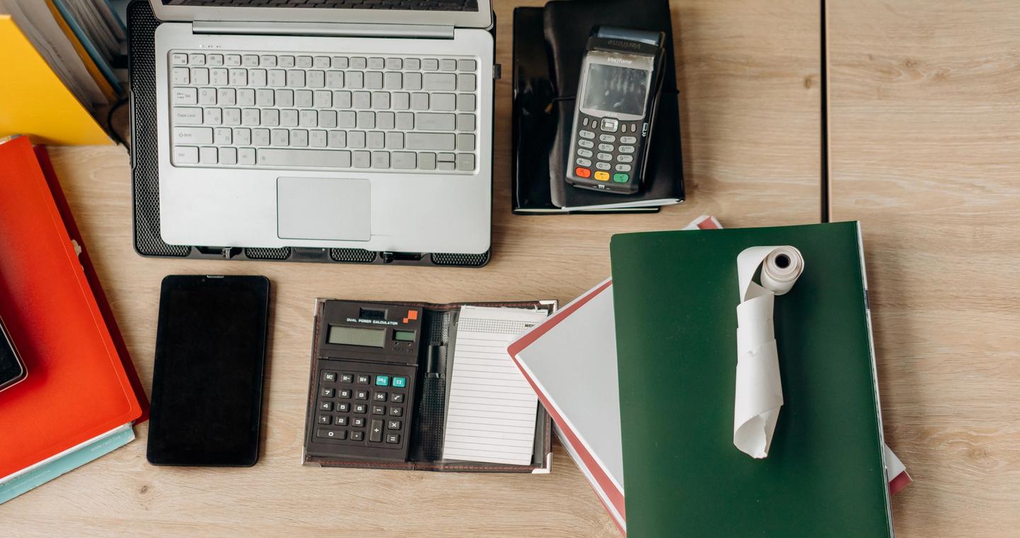 A desk holds a laptop, calculators, a payment terminal and notebooks.