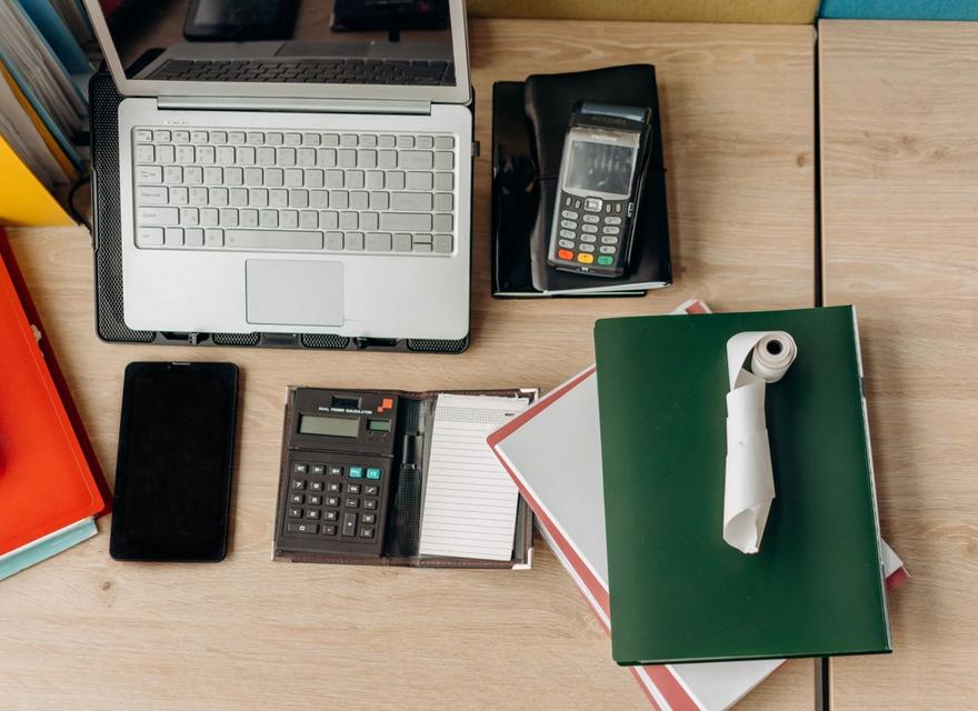 A desk holds a laptop, calculators, a payment terminal and notebooks.