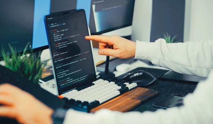 A person points at code on a laptop beside a monitor.