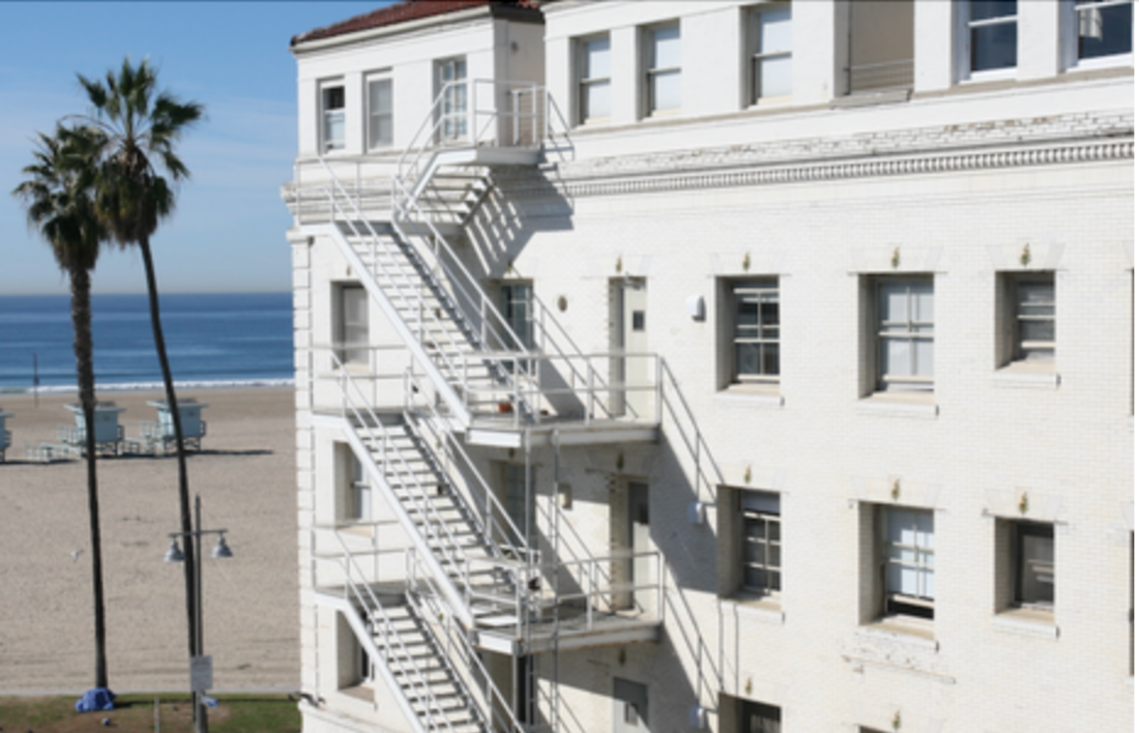 Side view of the former Waldorf Hotel, an early 20th century white building, with a fire escape. Venice Beach is in the background.