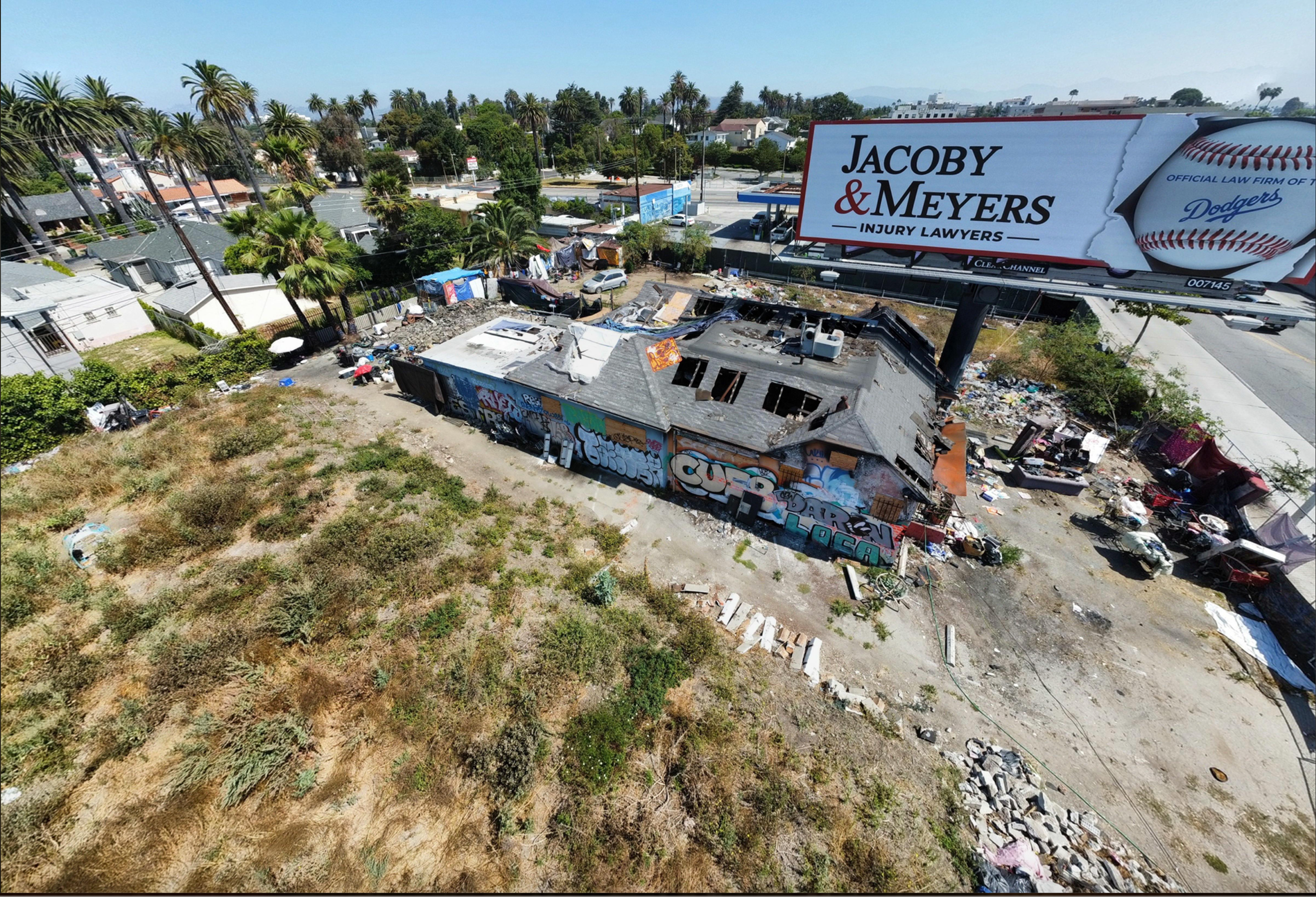 Drone shot of 1025 S. Crenshaw Blvd., a one-story commercial property, showing holes in the roof, heavy tagging, trash, and overgrown weeds.