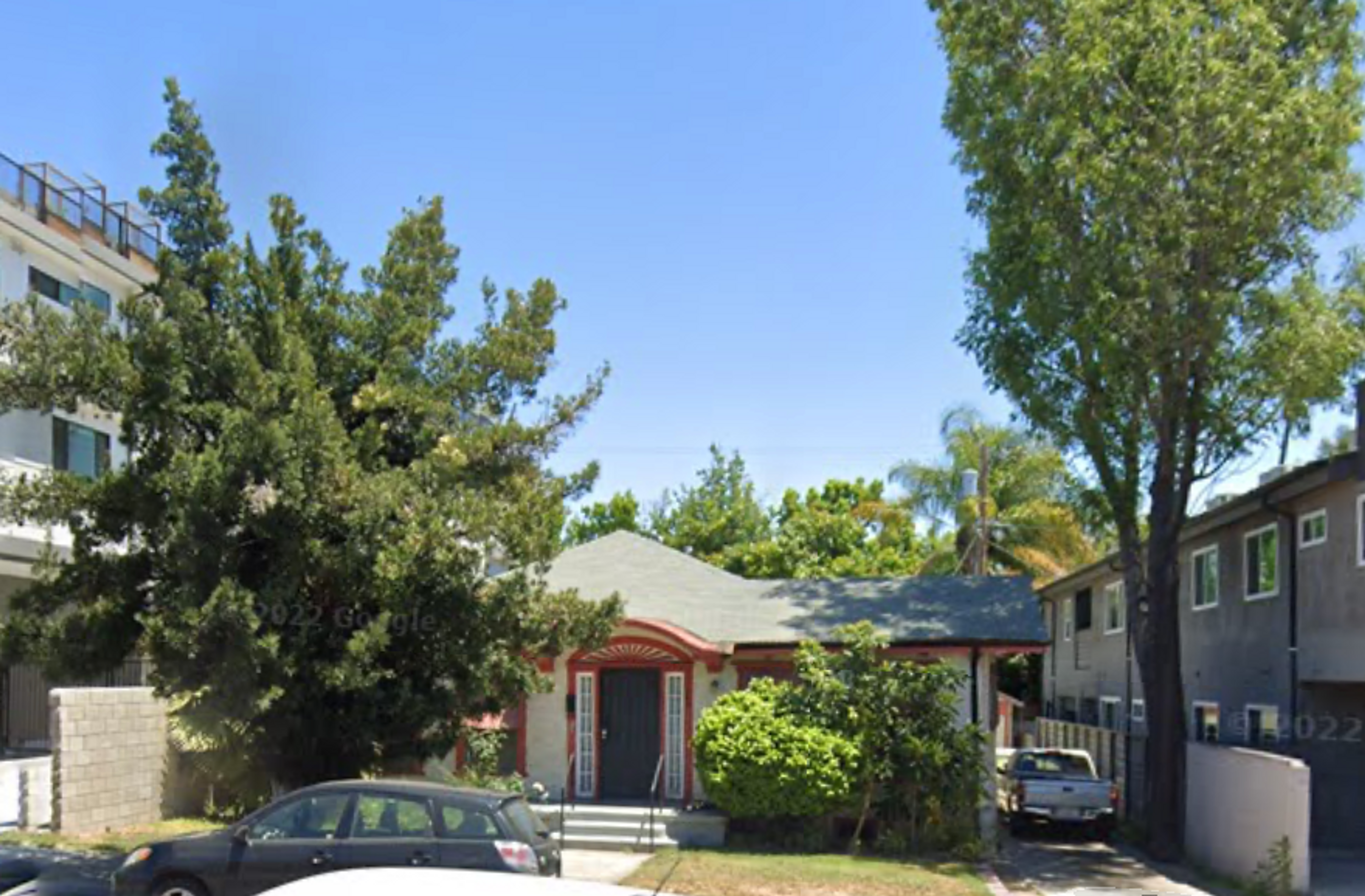1346 North Fairfax Avenue, a 1919 bungalow flanked by mature trees and two taller buildings. The house has burned down. Image courtesy of Google Maps.