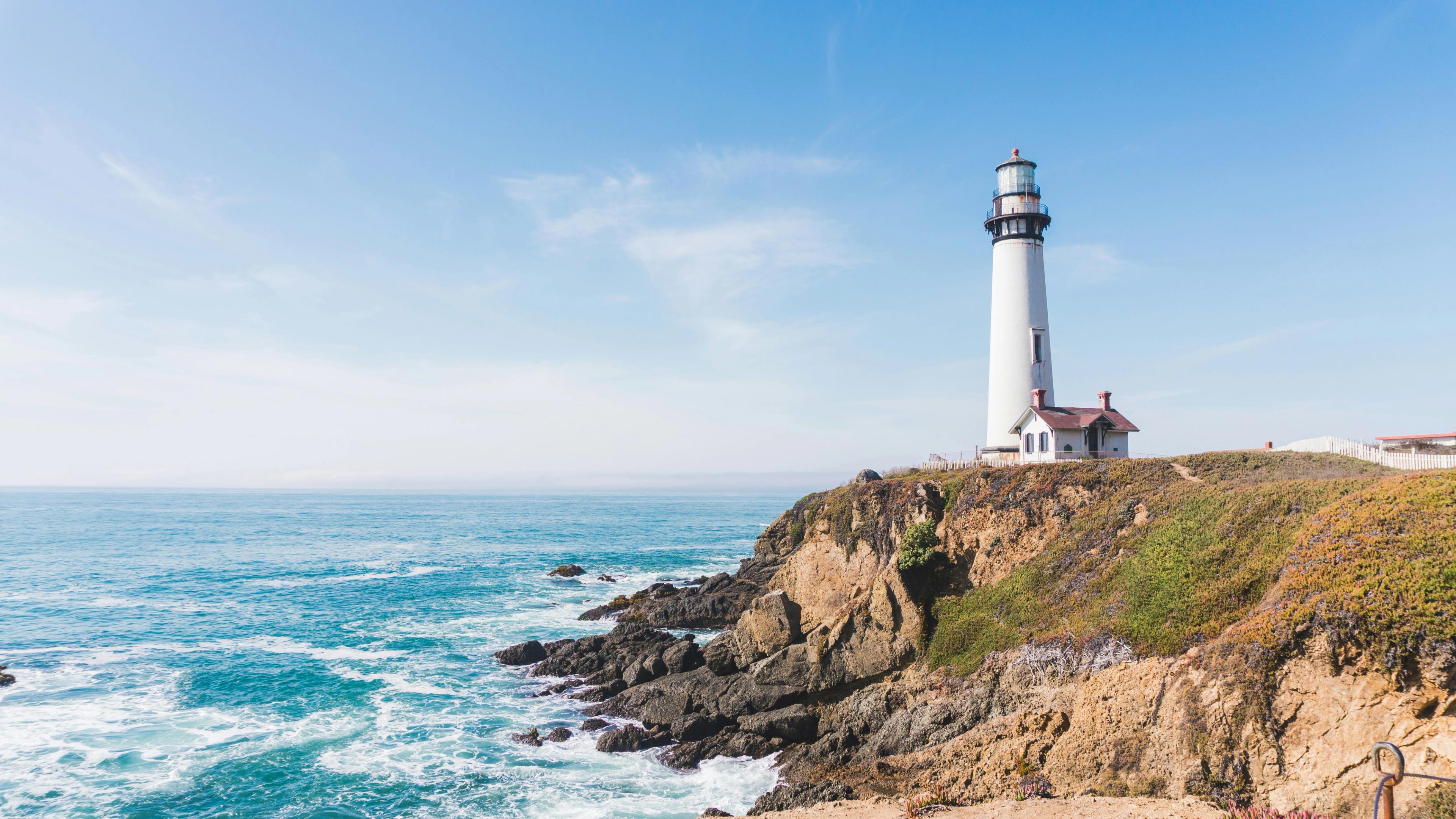 A white lighthouse and keeper's cottage stand on a rocky coastal bluff as waves break on the rocks below under a clear blue sky.