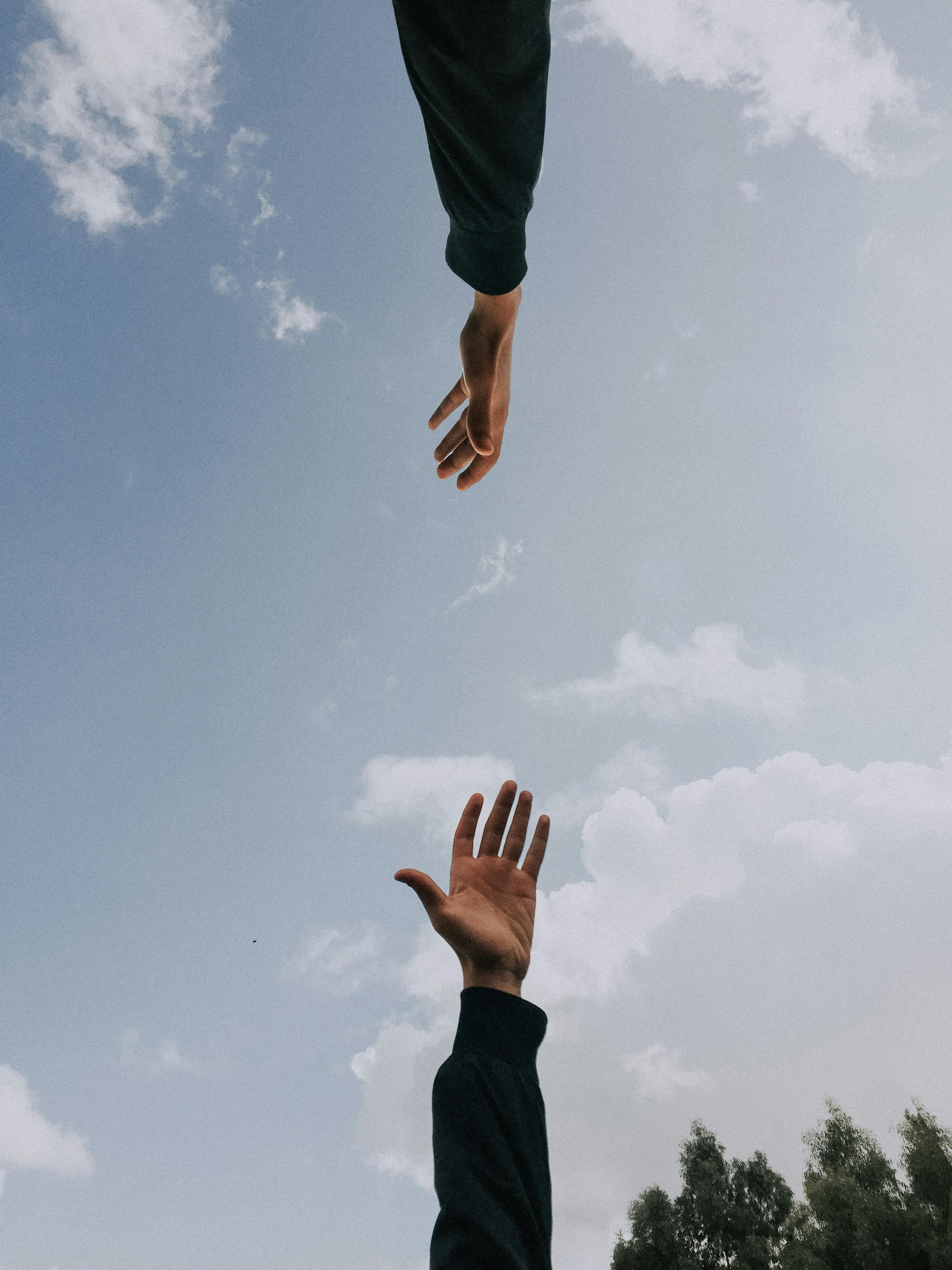 Two arms in dark sleeves reach toward each other against a blue sky with scattered clouds, one hand stretching down and the other up.