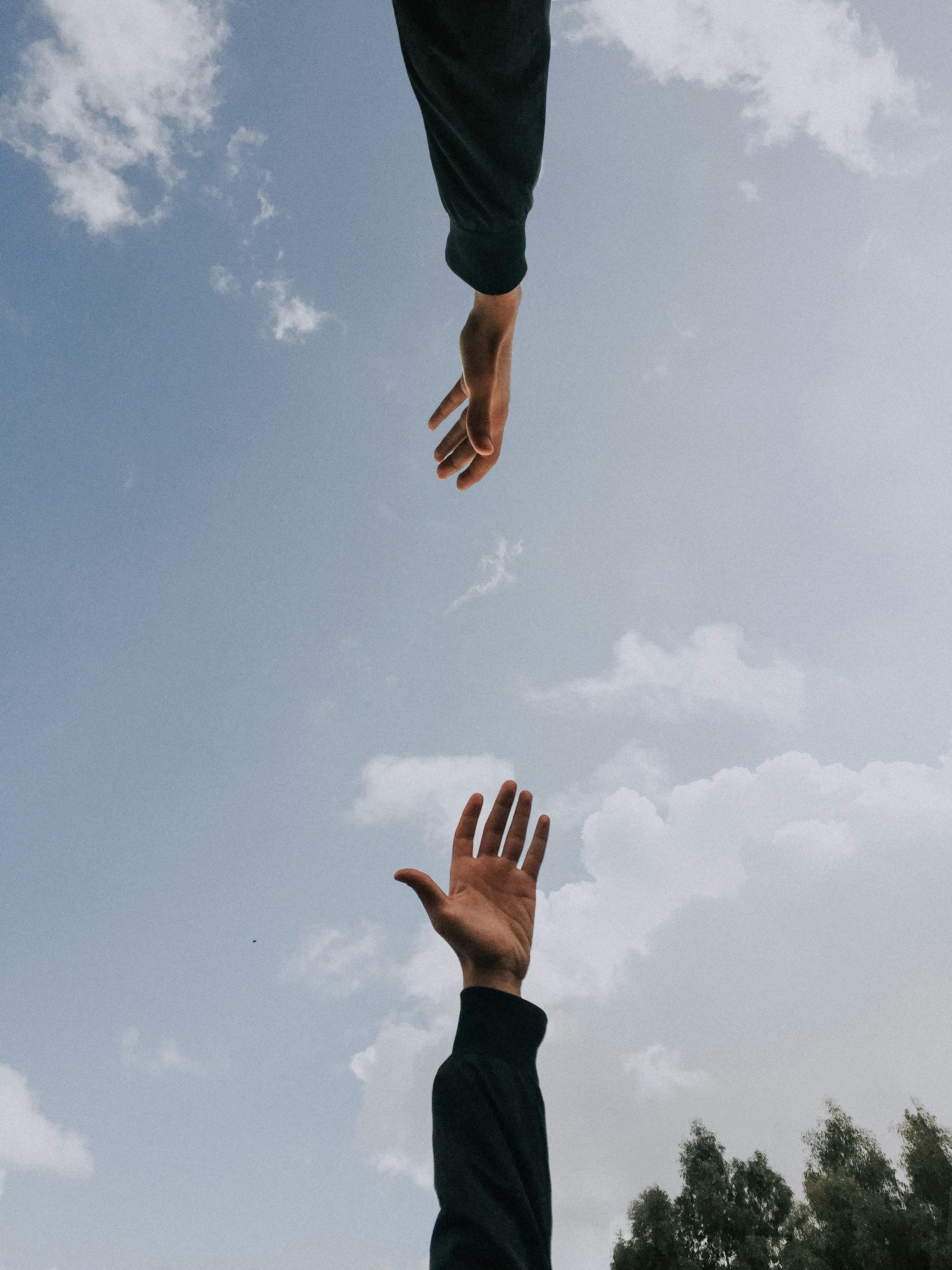Two arms in dark sleeves reach toward each other against a blue sky with scattered clouds, one hand stretching down and the other up.
