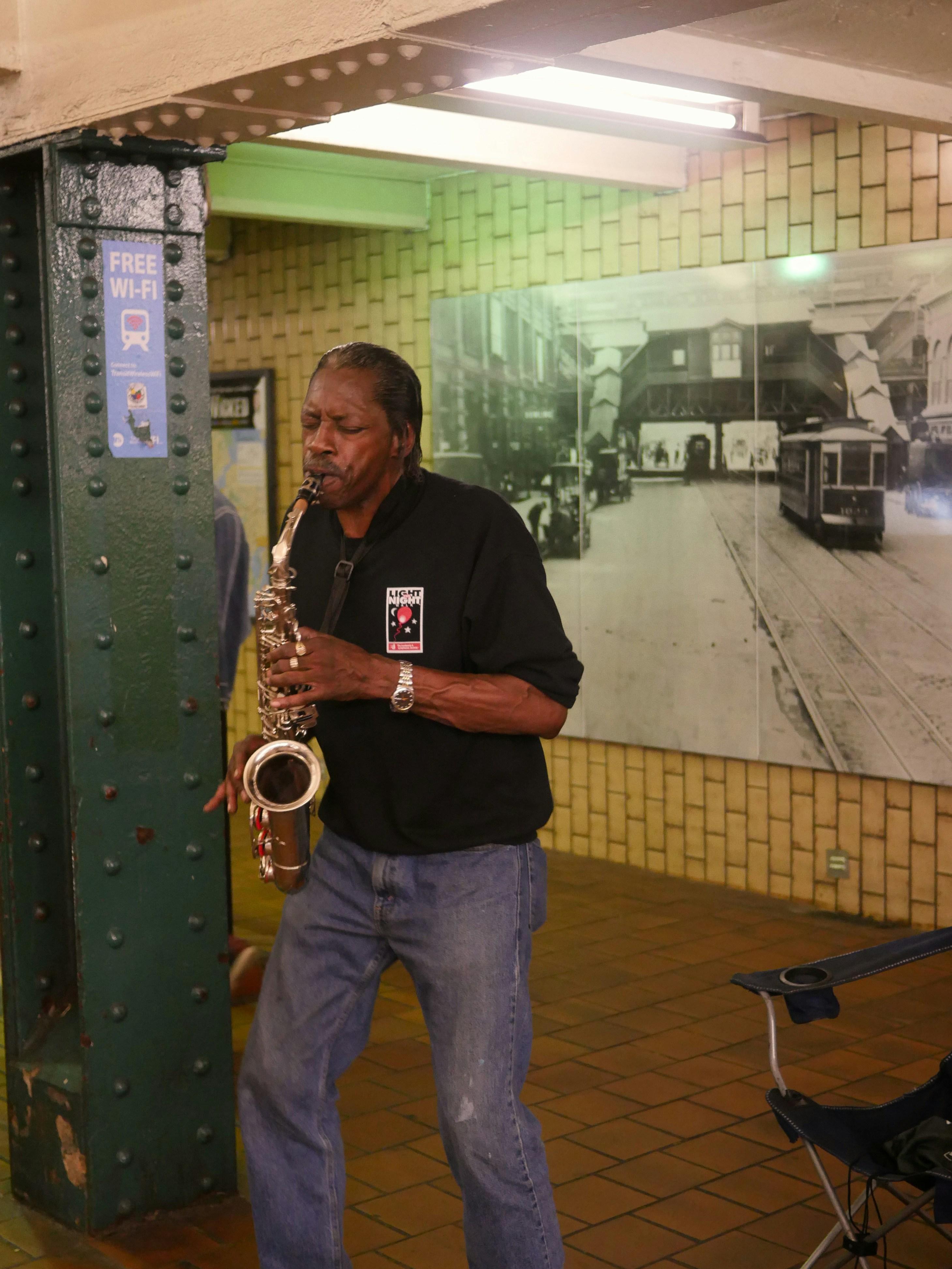 A man plays a saxophone beside a riveted green pillar in a tiled subway passage, in front of a large historic photograph of streetcars.