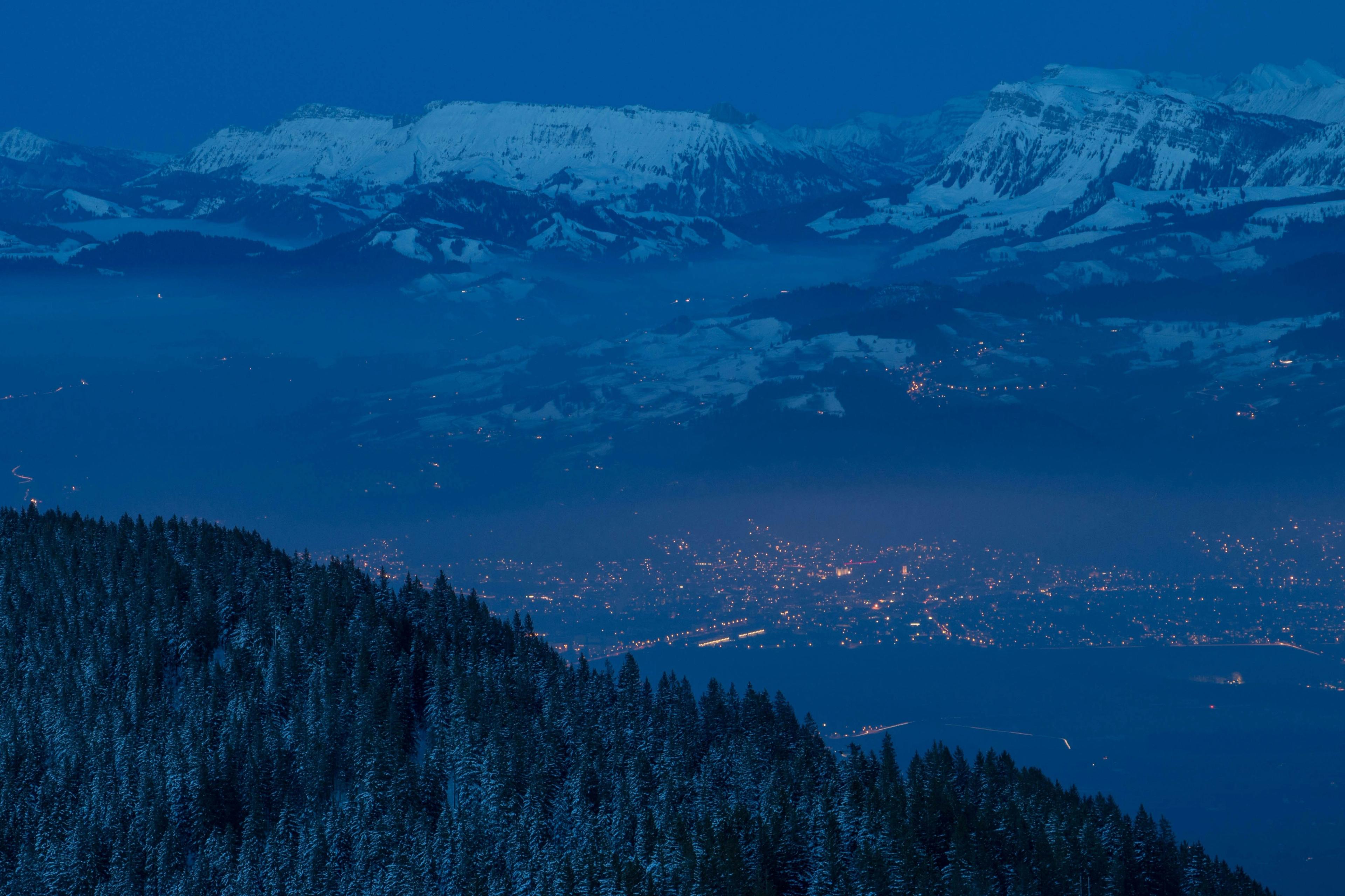A valley town glows with lights below snow-covered alpine peaks in blue evening haze, with a forested slope in the foreground.
