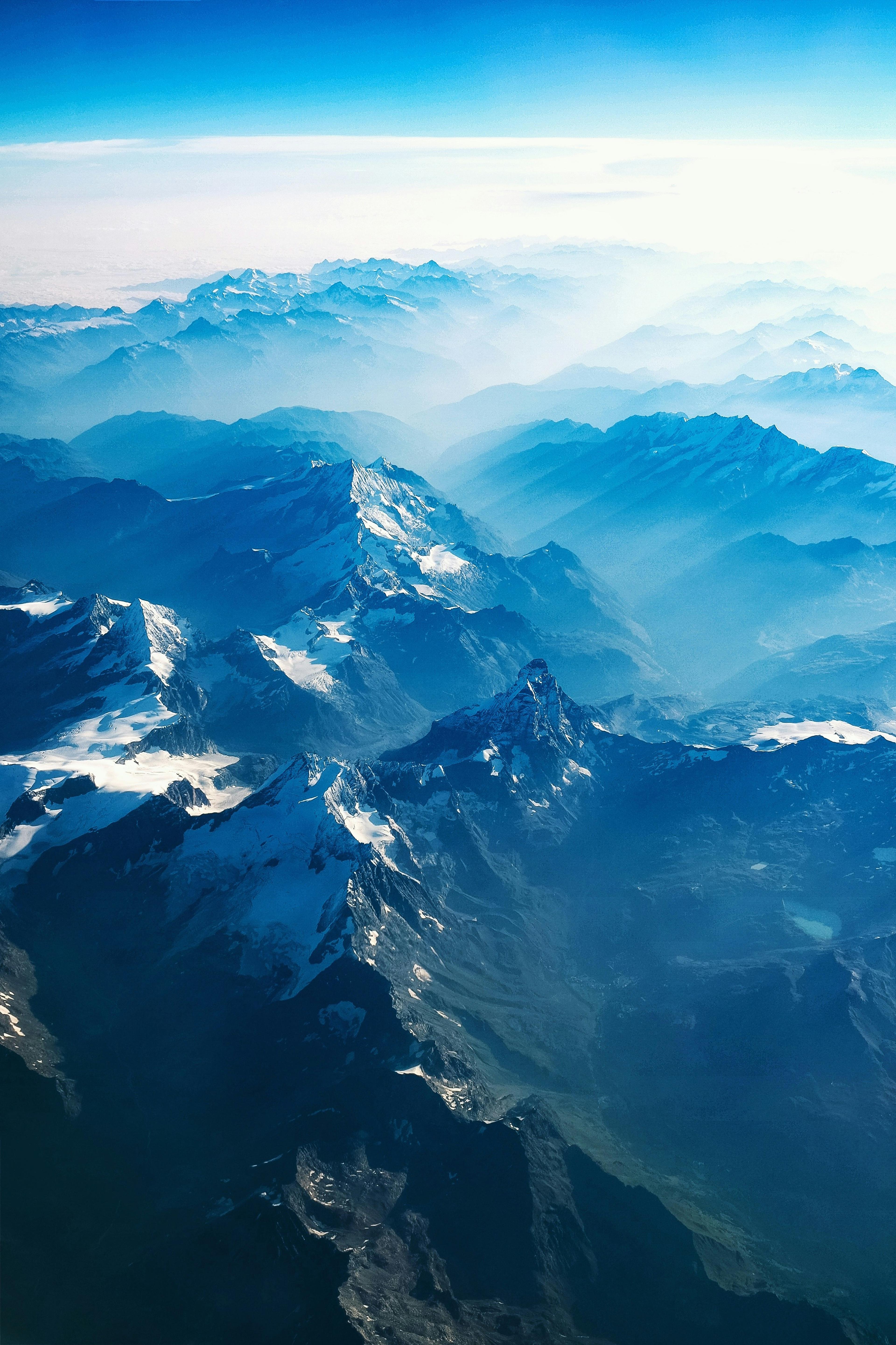 Aerial view of snow-capped mountain ridges receding into blue haze under a clear sky.