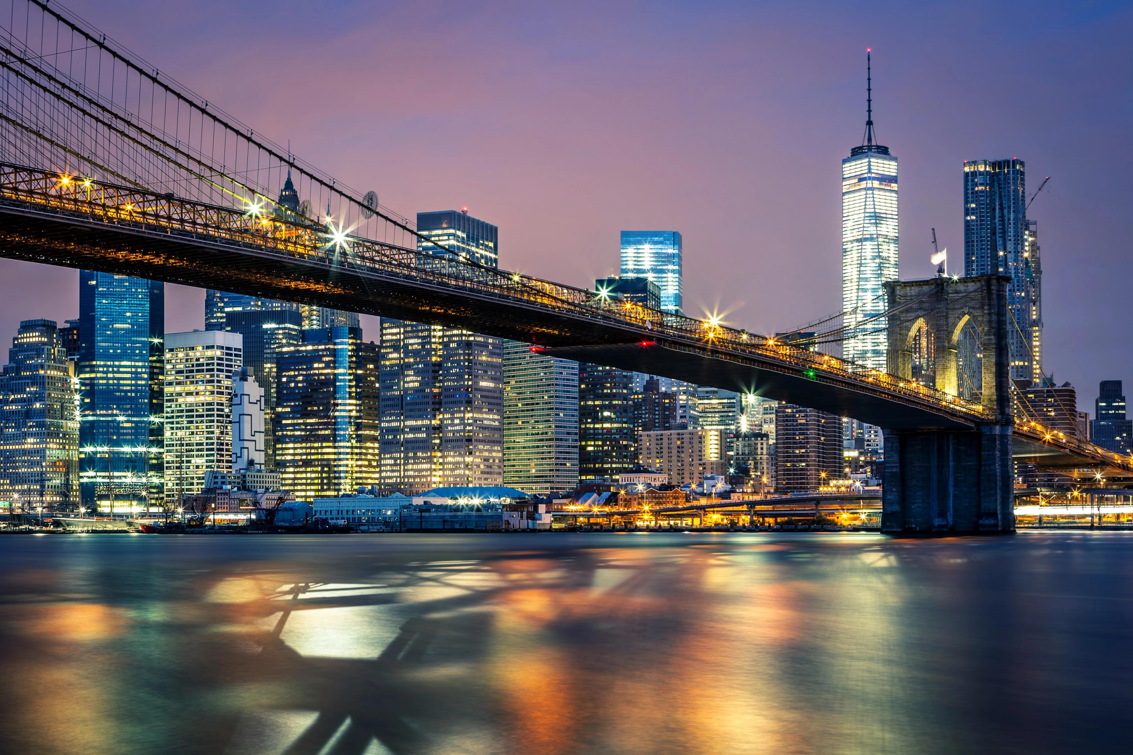 The Brooklyn Bridge stretches across the East River toward the lit skyscrapers of Lower Manhattan at dusk, their lights reflecting in the water.