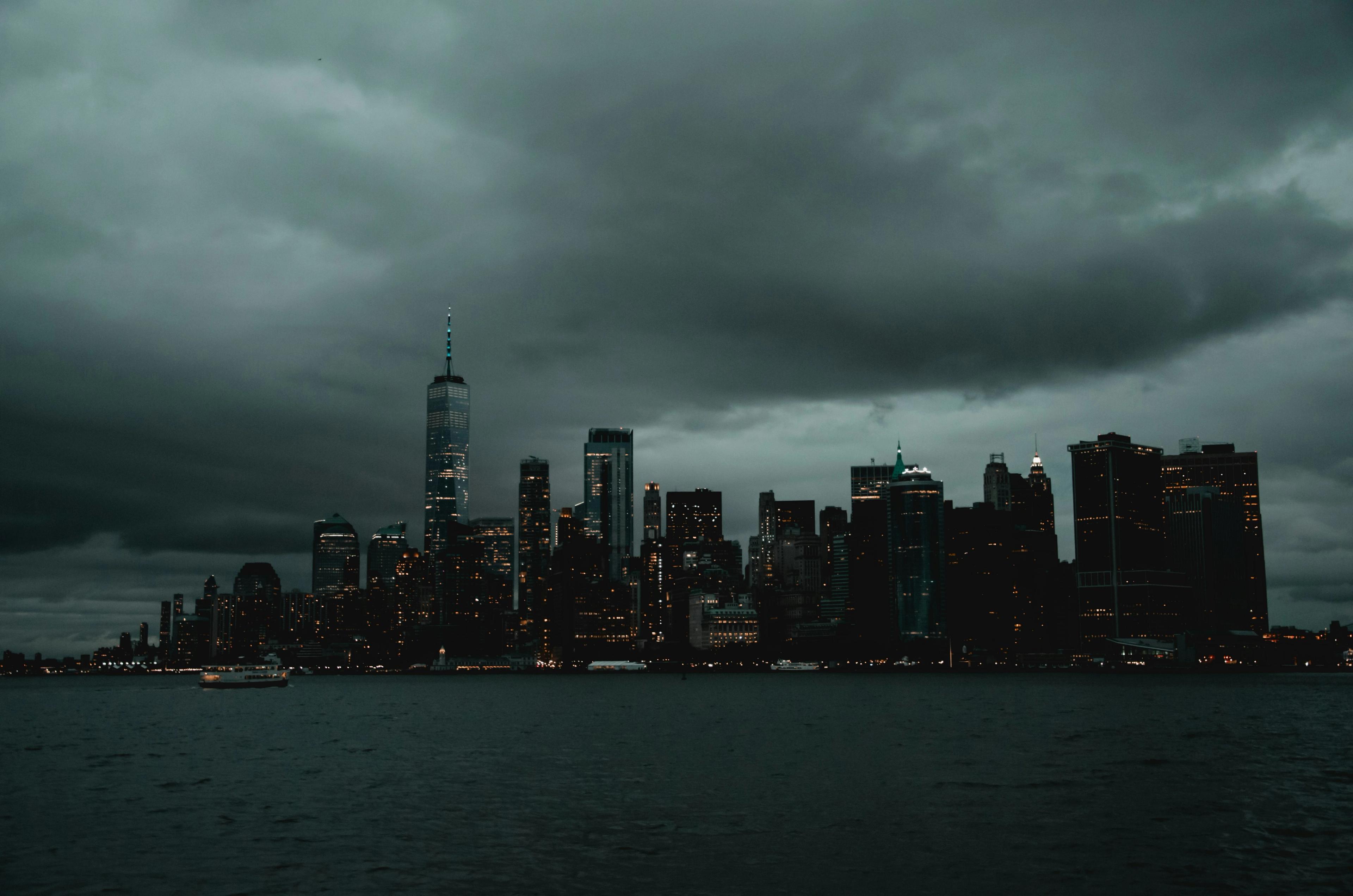 The Lower Manhattan skyline, dominated by One World Trade Center, stands dimly lit under heavy storm clouds with a ferry crossing the dark harbor.