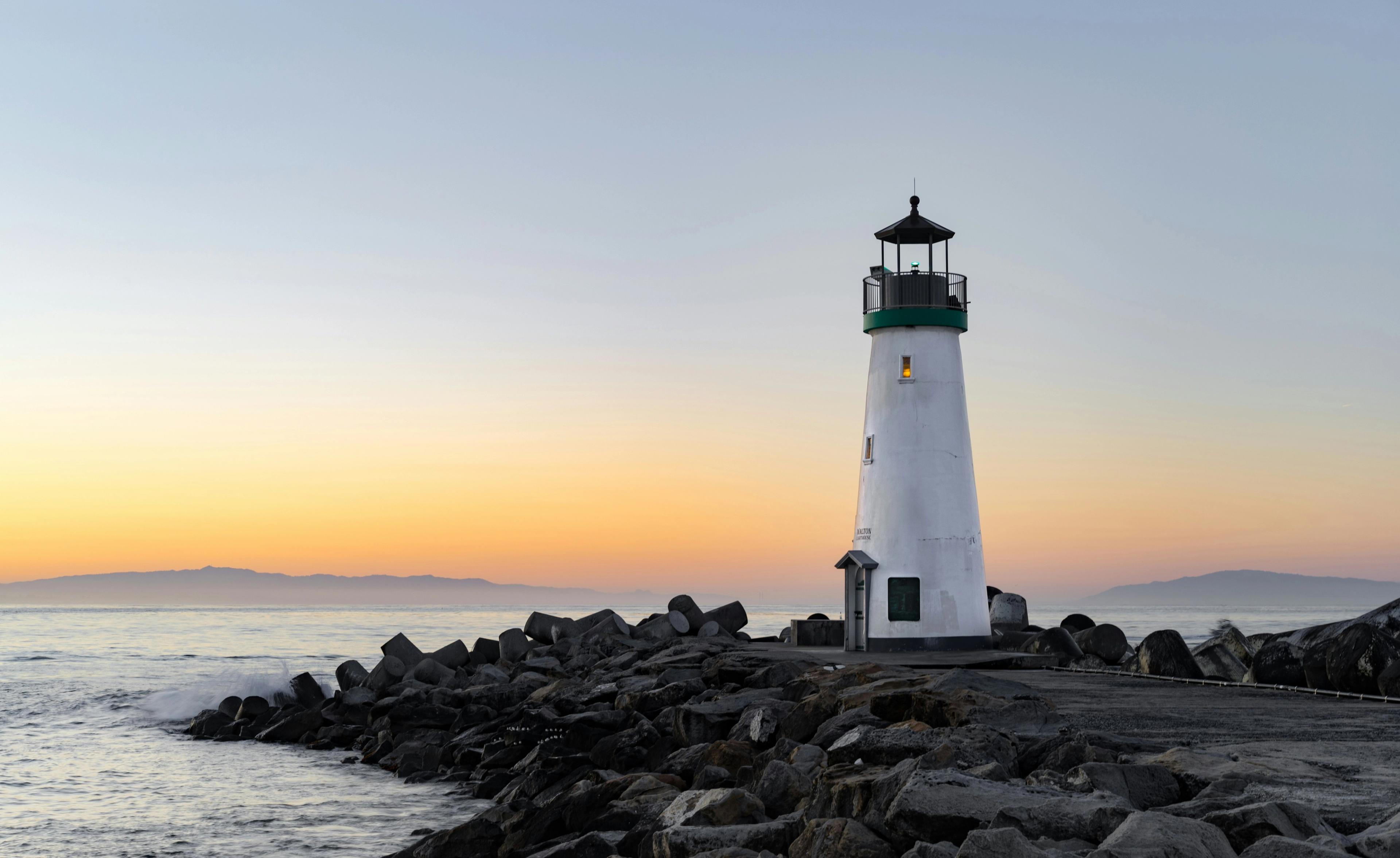 A small white lighthouse with a green trim band stands on a rocky breakwater at dusk, with an orange glow on the horizon behind it.
