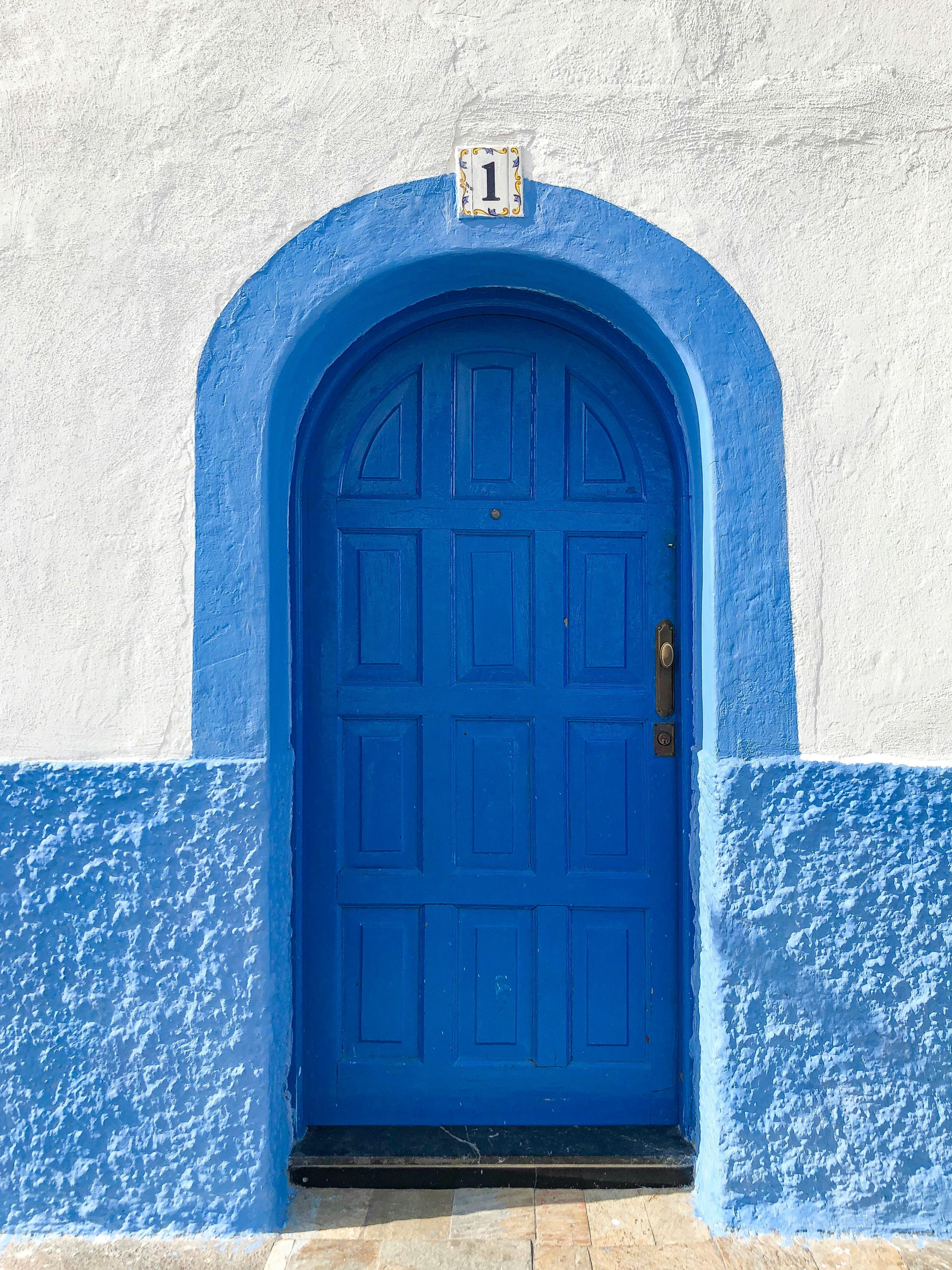 An arched blue wooden door numbered 1 set into a whitewashed wall with a blue-painted lower band.