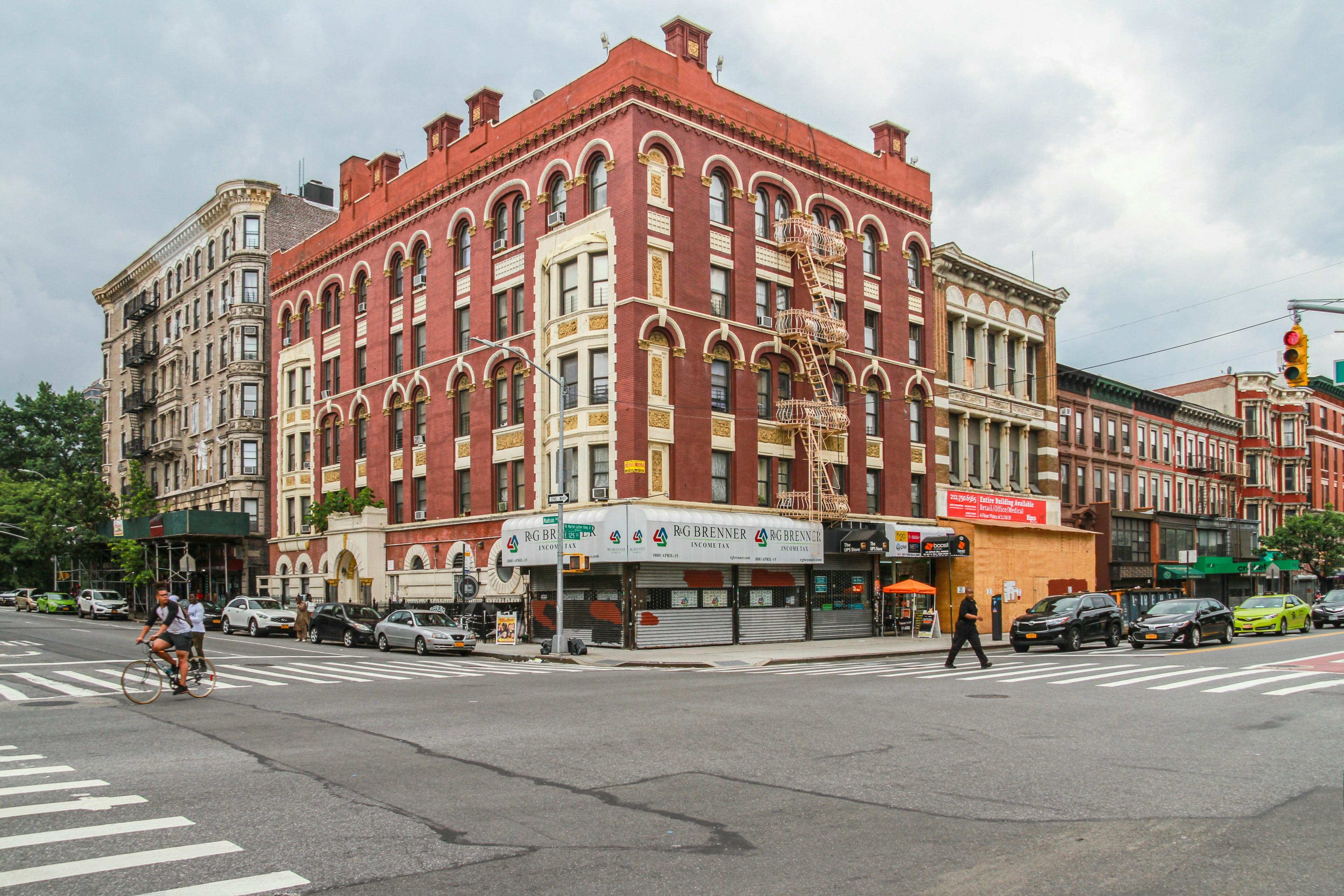 A cyclist and pedestrians cross a wide intersection in front of a red-brick corner building with fire escapes and shuttered storefronts on East 125th Street.