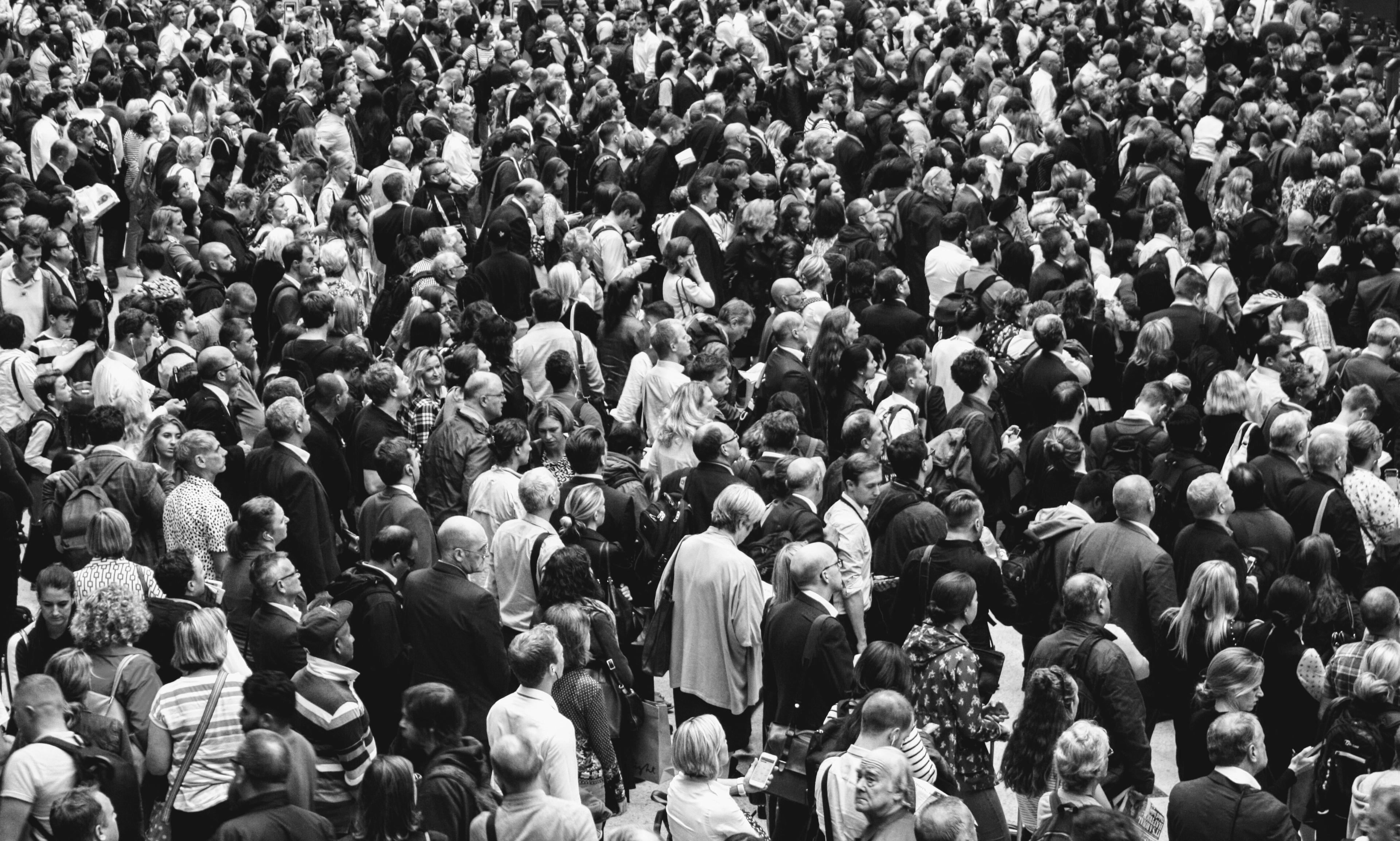A dense crowd of commuters seen from above fills a station concourse in black and white, most facing away toward departure boards.