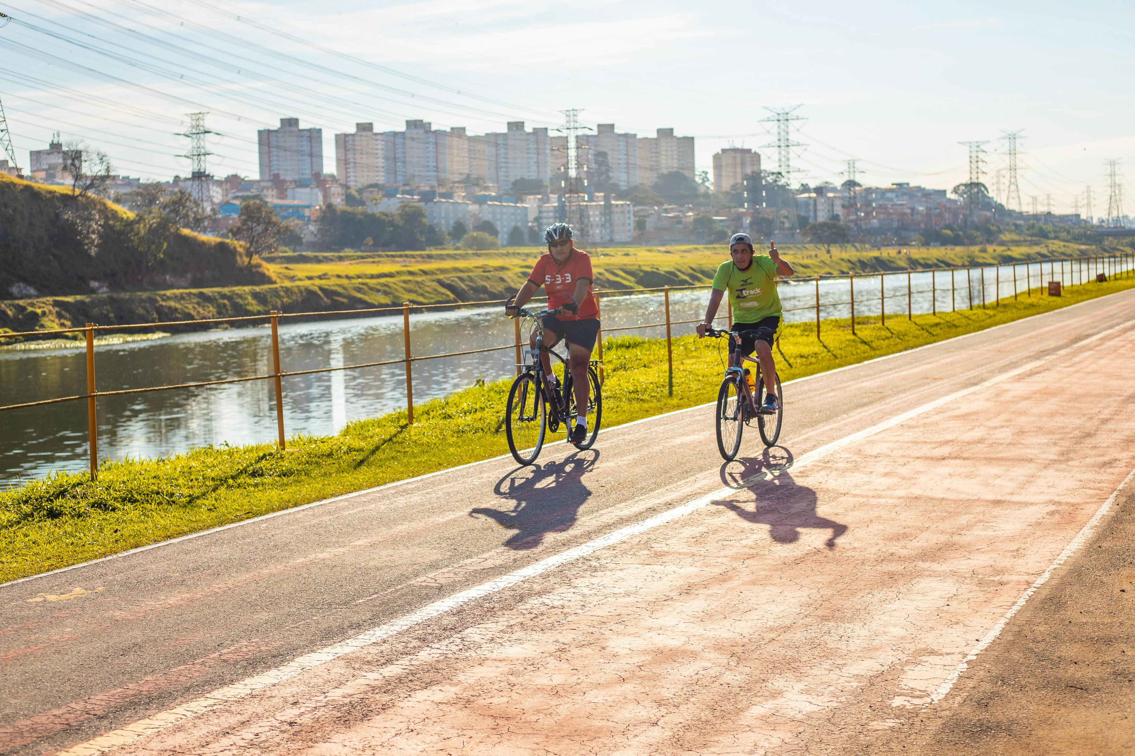 Two men ride bicycles along a riverside cycle path in morning light, with a fence, a river, apartment blocks and power pylons behind them.