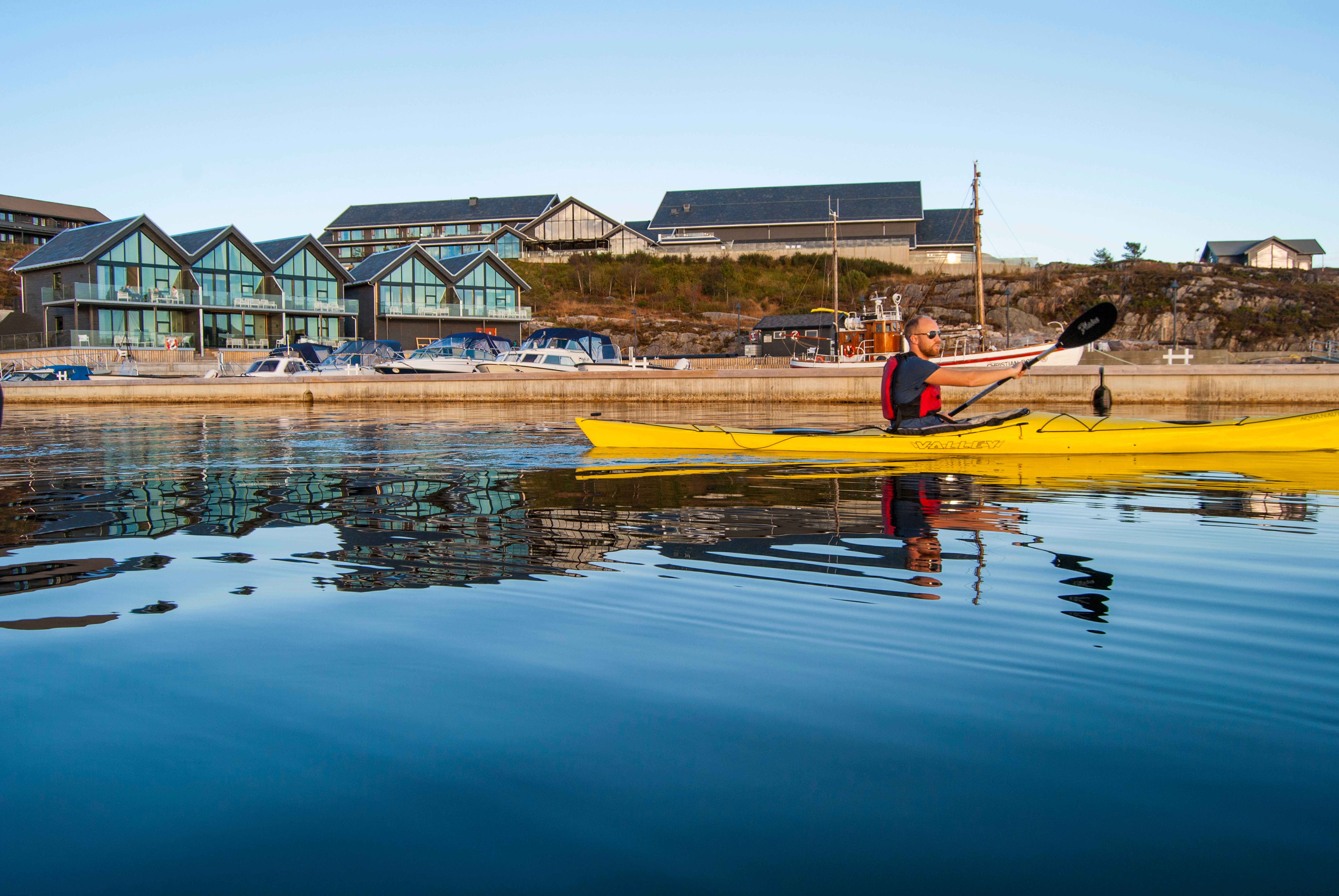 A guy kayaking on a perfectly calm sea, with Panorama hotel and marina in the background