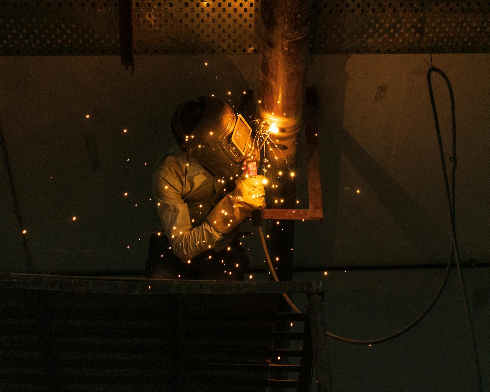 A welder at work indoors, creating bright sparks while welding metal pipes.
