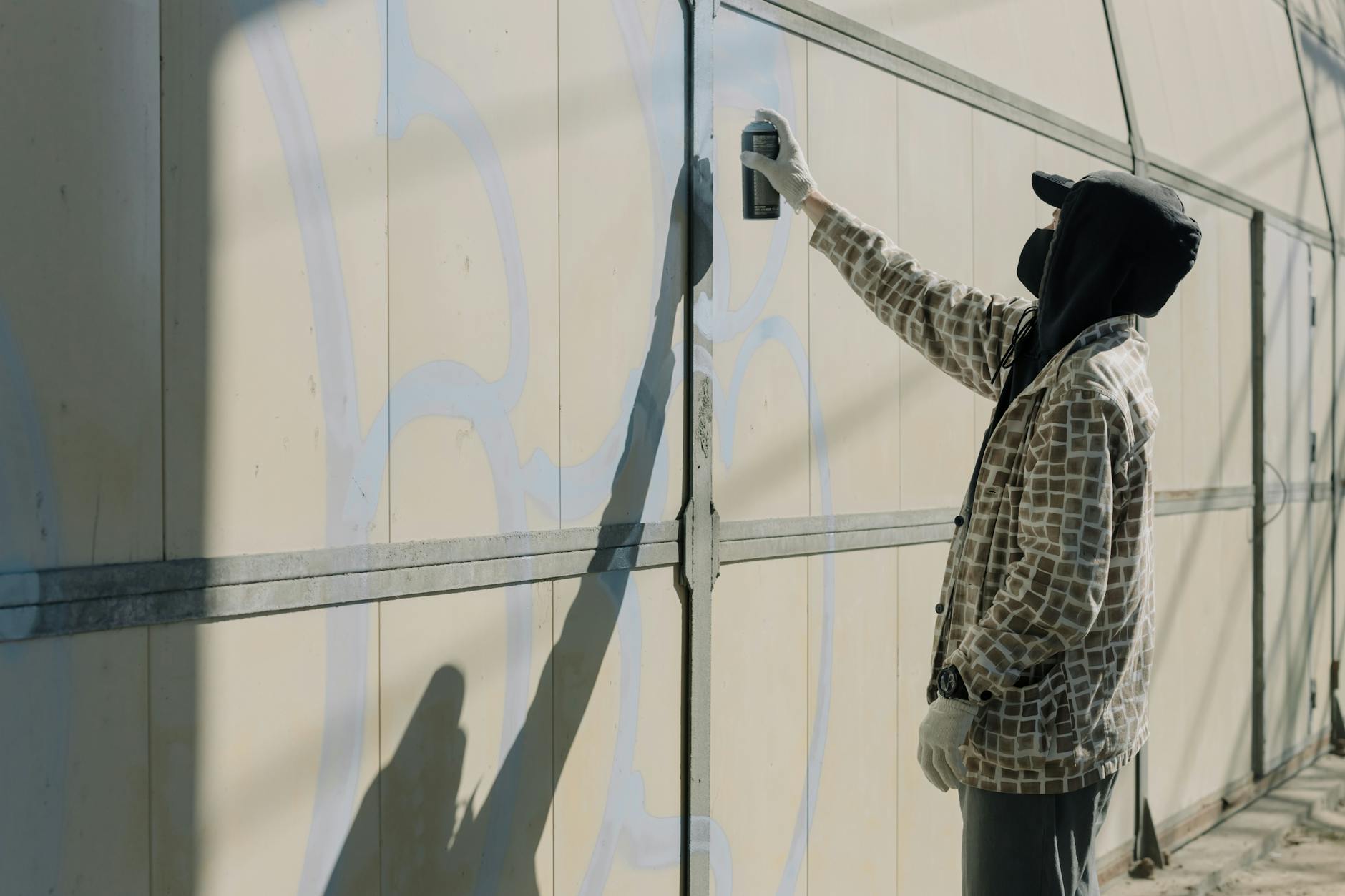 A person in a hoodie spray painting graffiti on an outdoor wall in daylight.