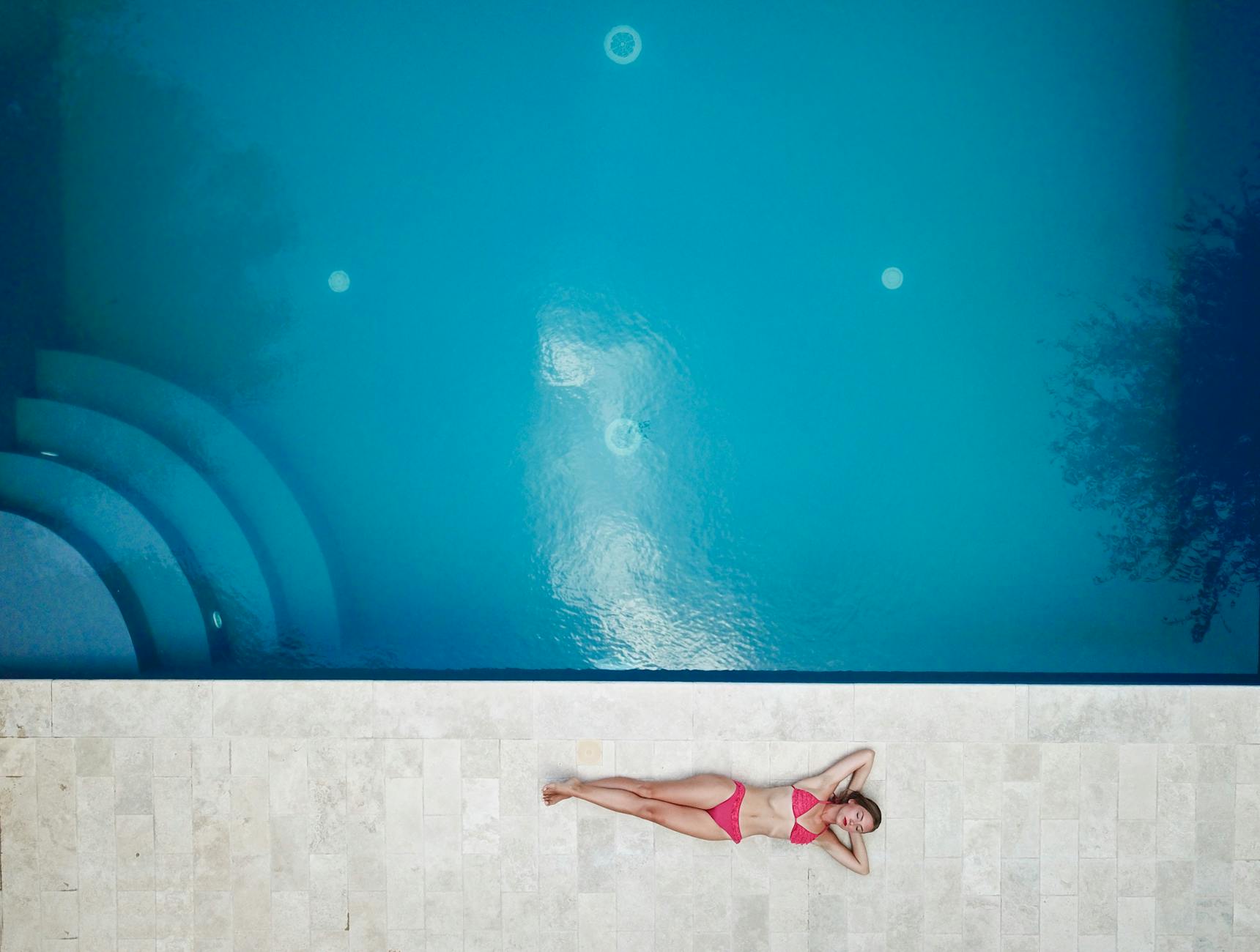 Bird's-eye view of a woman in a pink bikini relaxing by a bright blue swimming pool.
