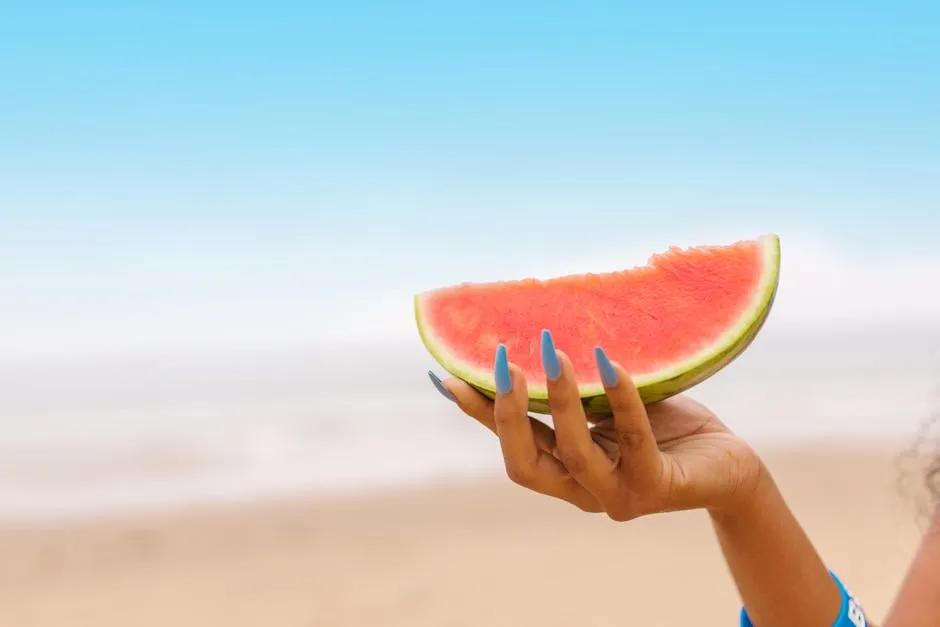Hand med blå sommarnaglar håller i en vattenmelonbit en solig dag