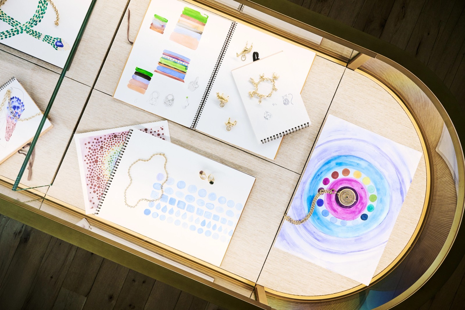 Overhead view of a display case with jewelry design sketchbooks, color swatches, and gold jewelry pieces.