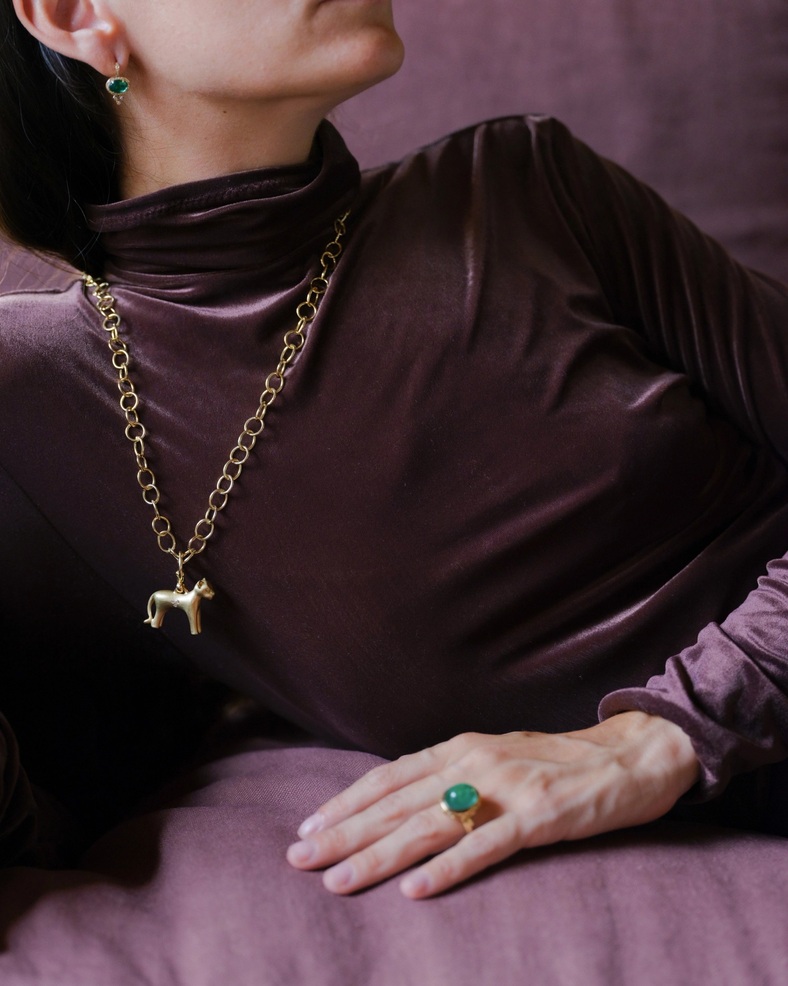 A person wears a plum velvet turtleneck, gold earrings with green stones, a gold chain with a dog pendant, and a matching green gemstone ring.