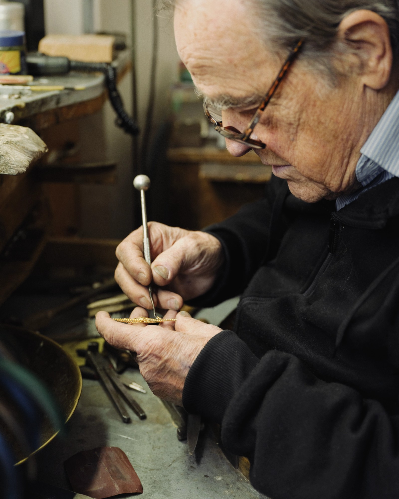 An older man with glasses meticulously works on a small gold piece with a tool at a workbench.