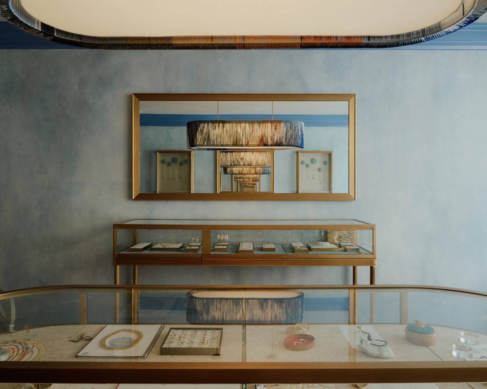 Elegant jewelry store with light blue walls, gold-framed jewelry display cases, and a mirror reflecting a fringed light fixture, creating an infinite reflection.