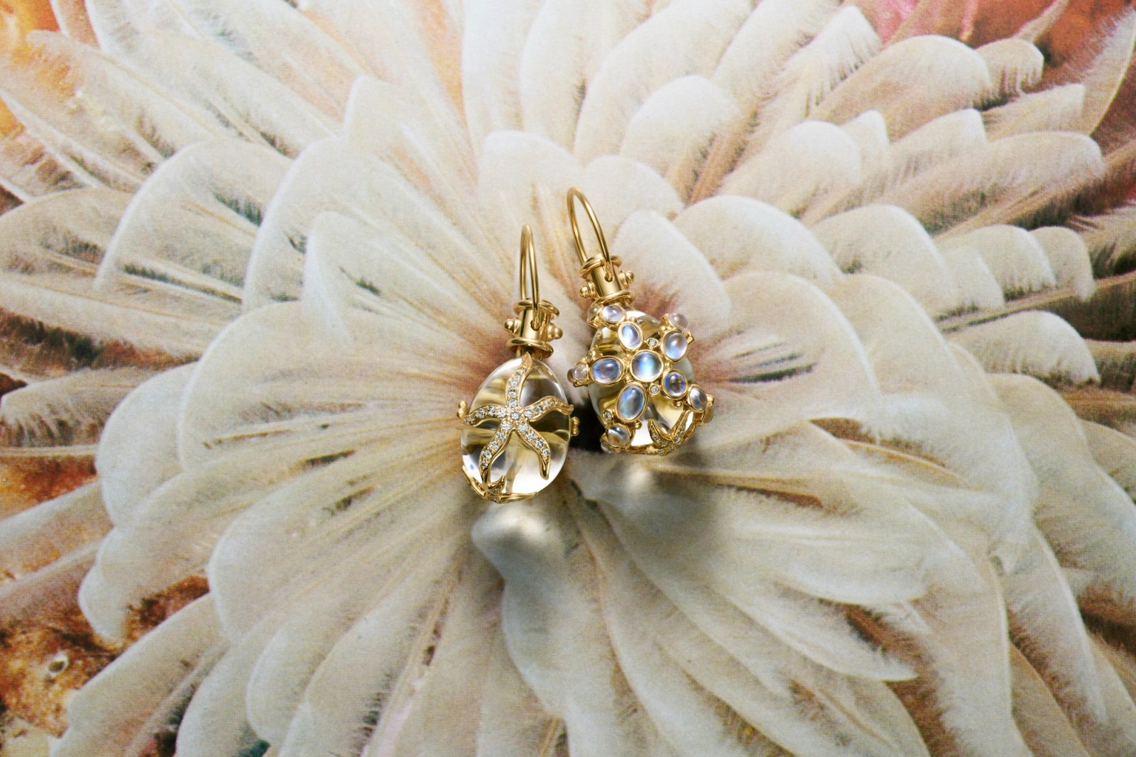 Gold drop earrings; one with a crystal starfish, the other with moonstones, on a light textured background.