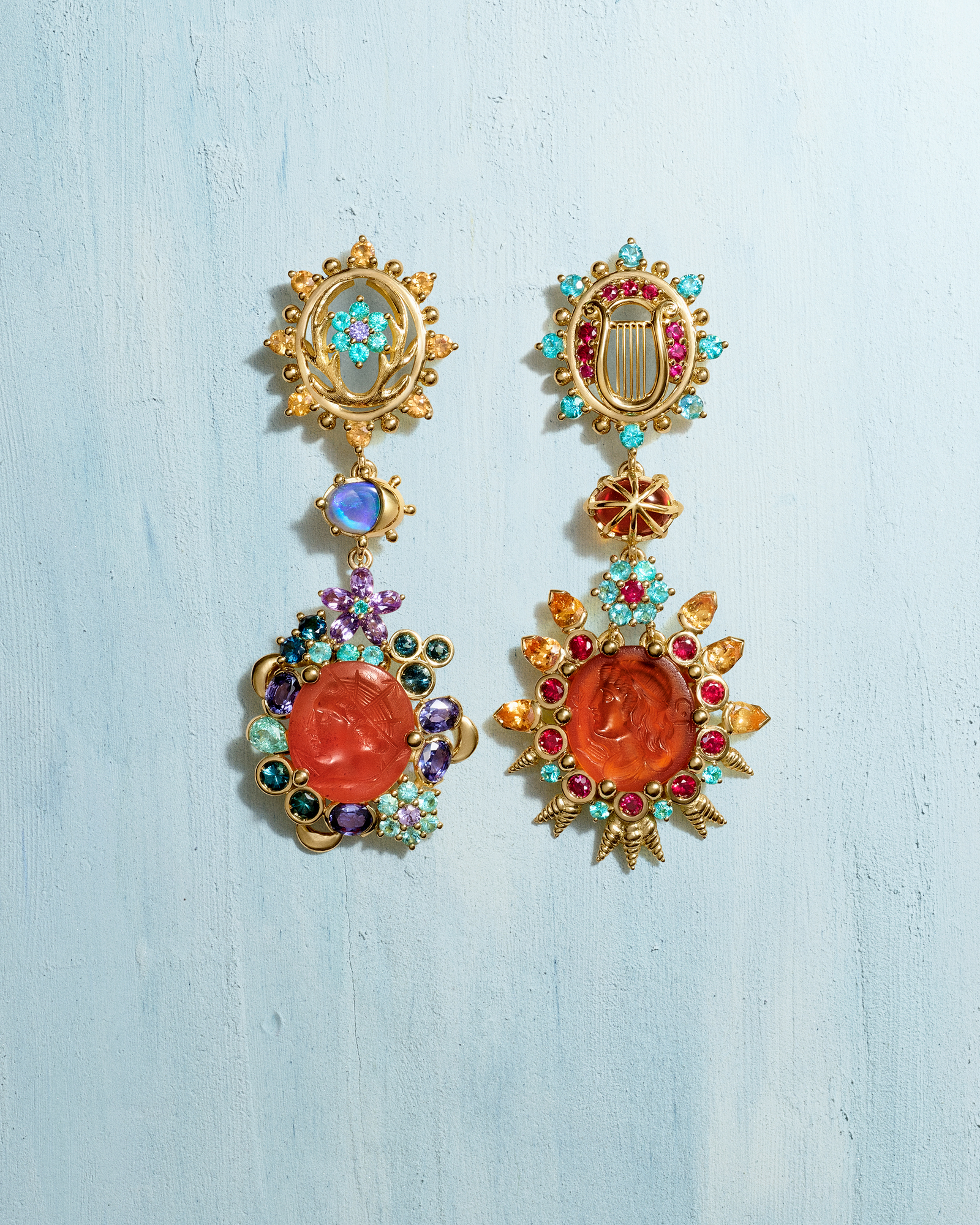 A pair of ornate, mismatched gold earrings with colorful gemstones and central carved orange stones, on a light blue textured surface.