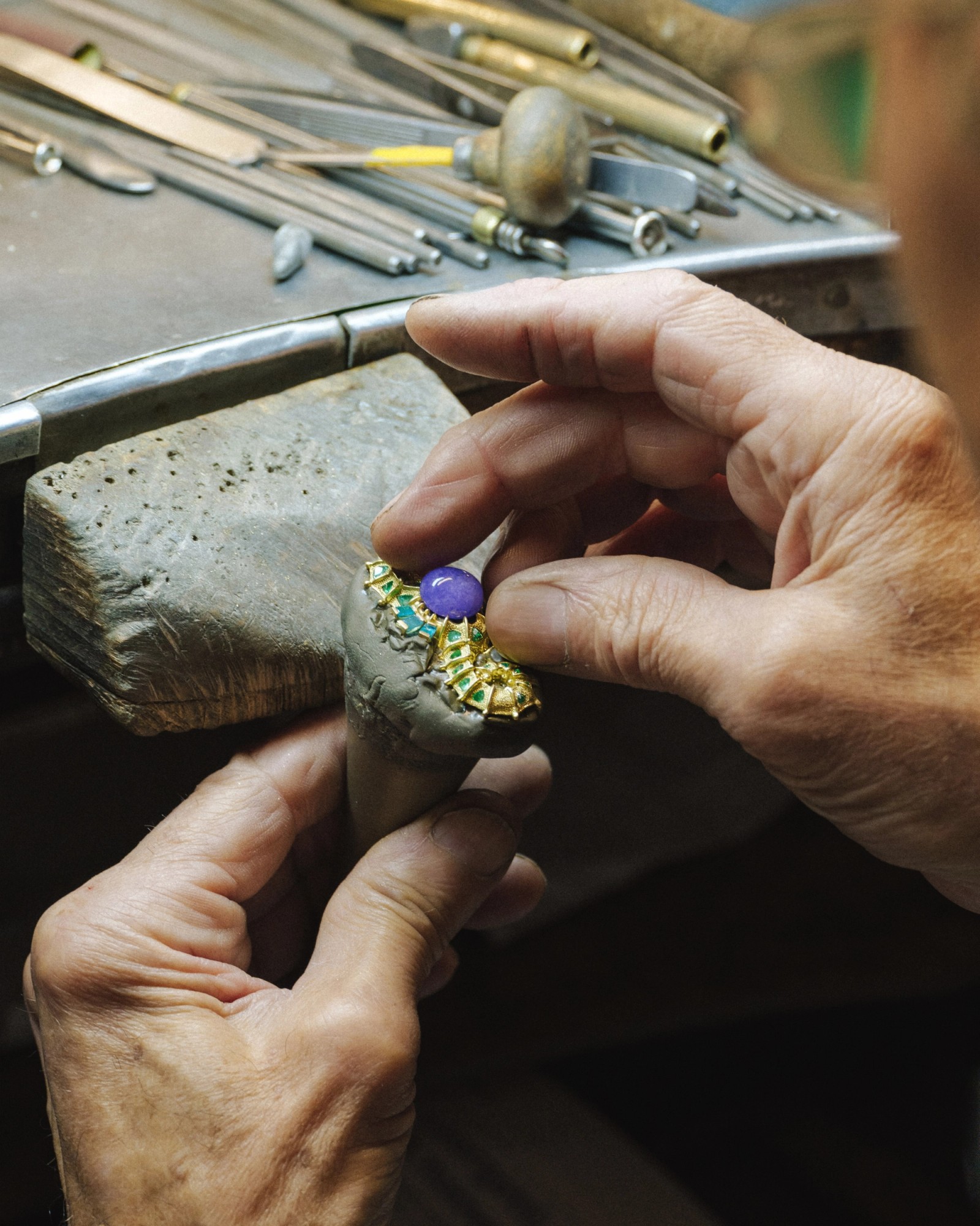 Hands setting a large purple gemstone and smaller green gemstones into an ornate gold jewelry piece on a workbench.