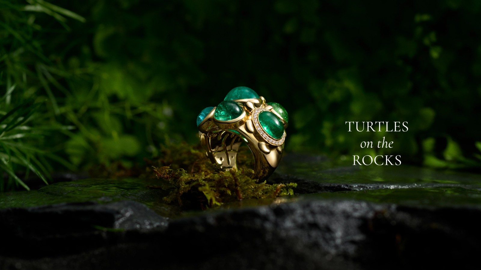 Gold ring with green emeralds and diamonds, designed as turtles, on mossy rocks. Text: 'TURTLES on the ROCKS'.