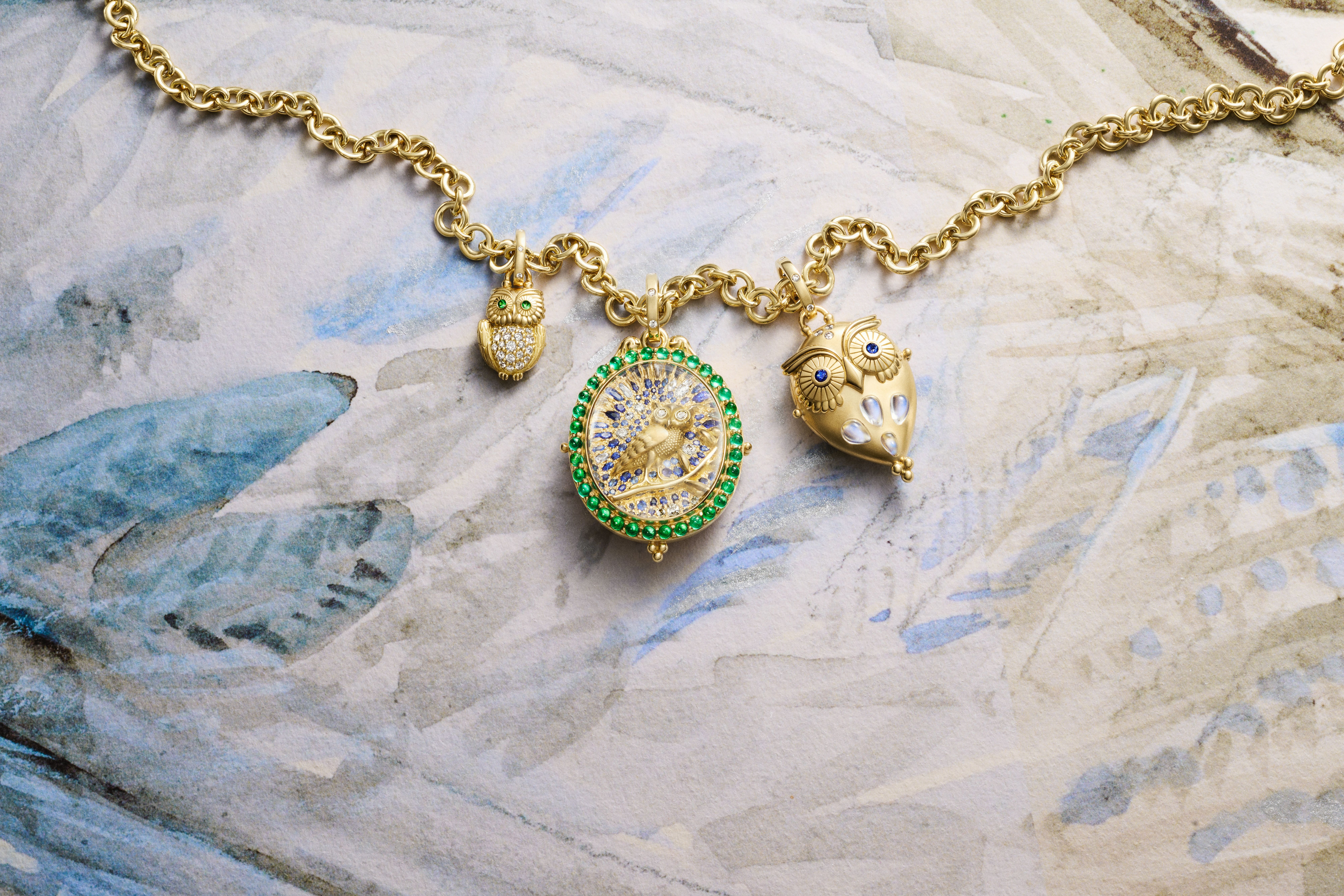 A gold necklace with three charms: a small diamond-bellied owl with green eyes, a round green-bordered bird pendant with blue and white gems, and a larger gold owl with blue eyes.